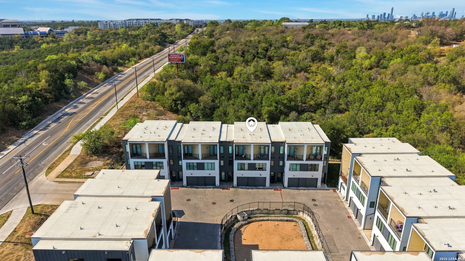 Aerial view of modern townhomes beside a wooded greenbelt, with a road and distant city skyline.