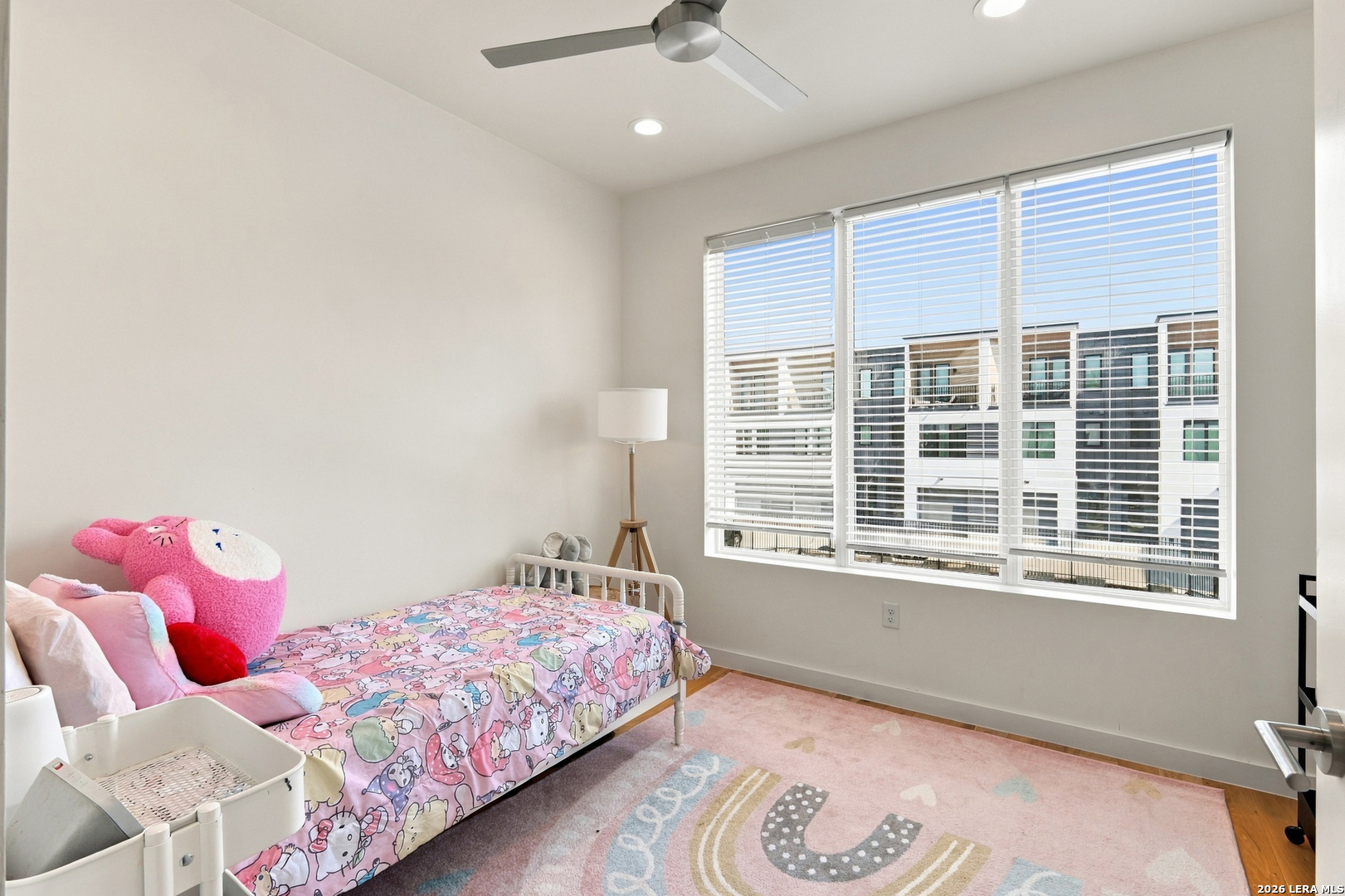 Furnished bedroom with pink bedding and rug, large windows, and views of neighboring townhomes.