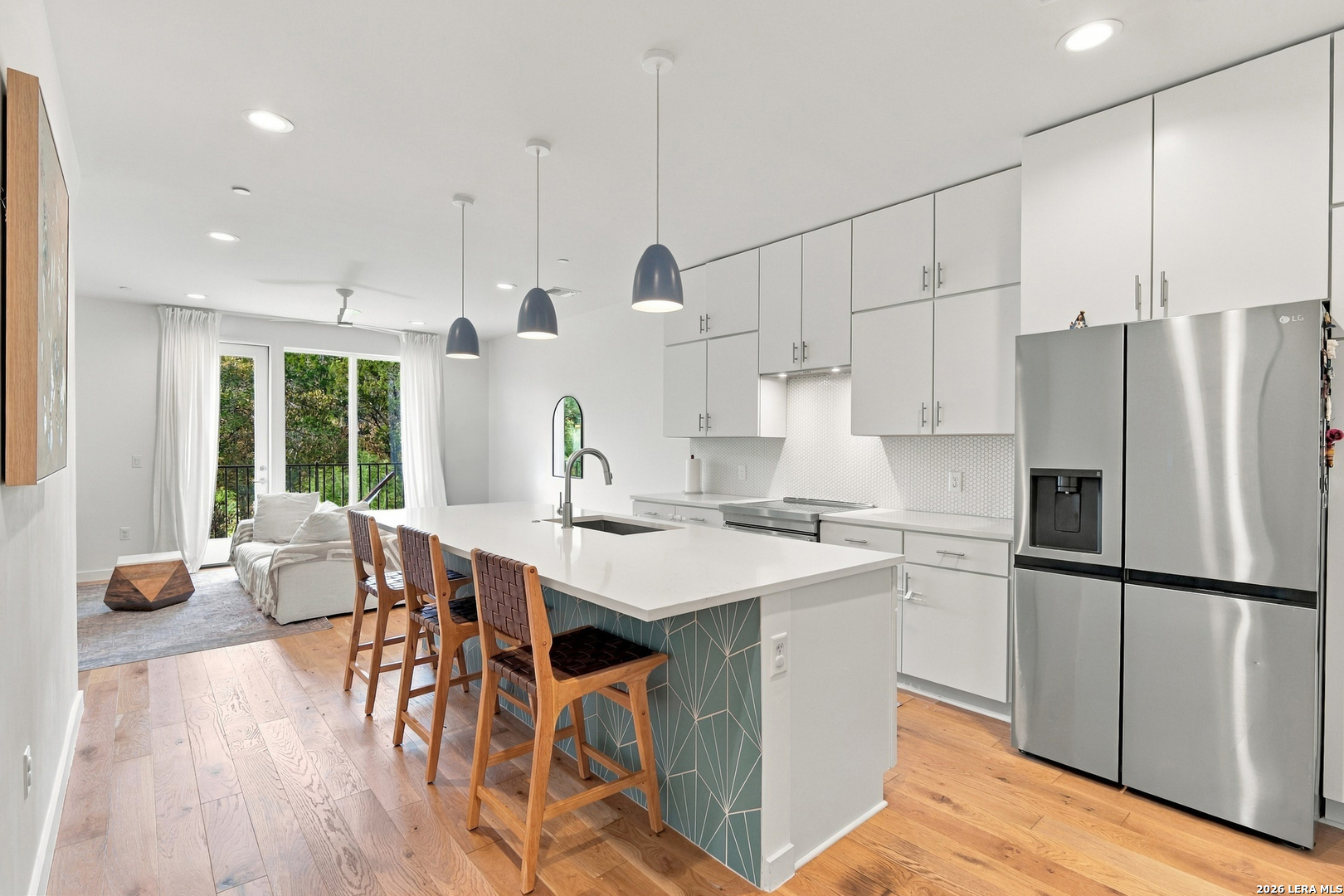 Open-plan kitchen with a teal island, wooden stools, stainless appliances, and sliding doors to trees.