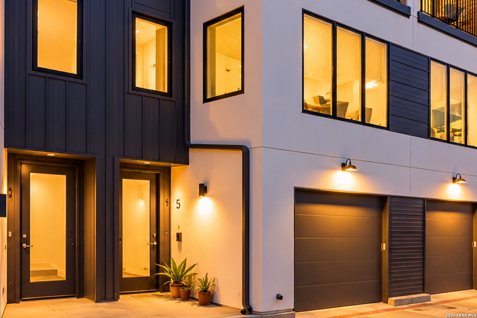 Modern townhouse exterior at dusk with glowing windows, glass entry doors, and two garage doors.