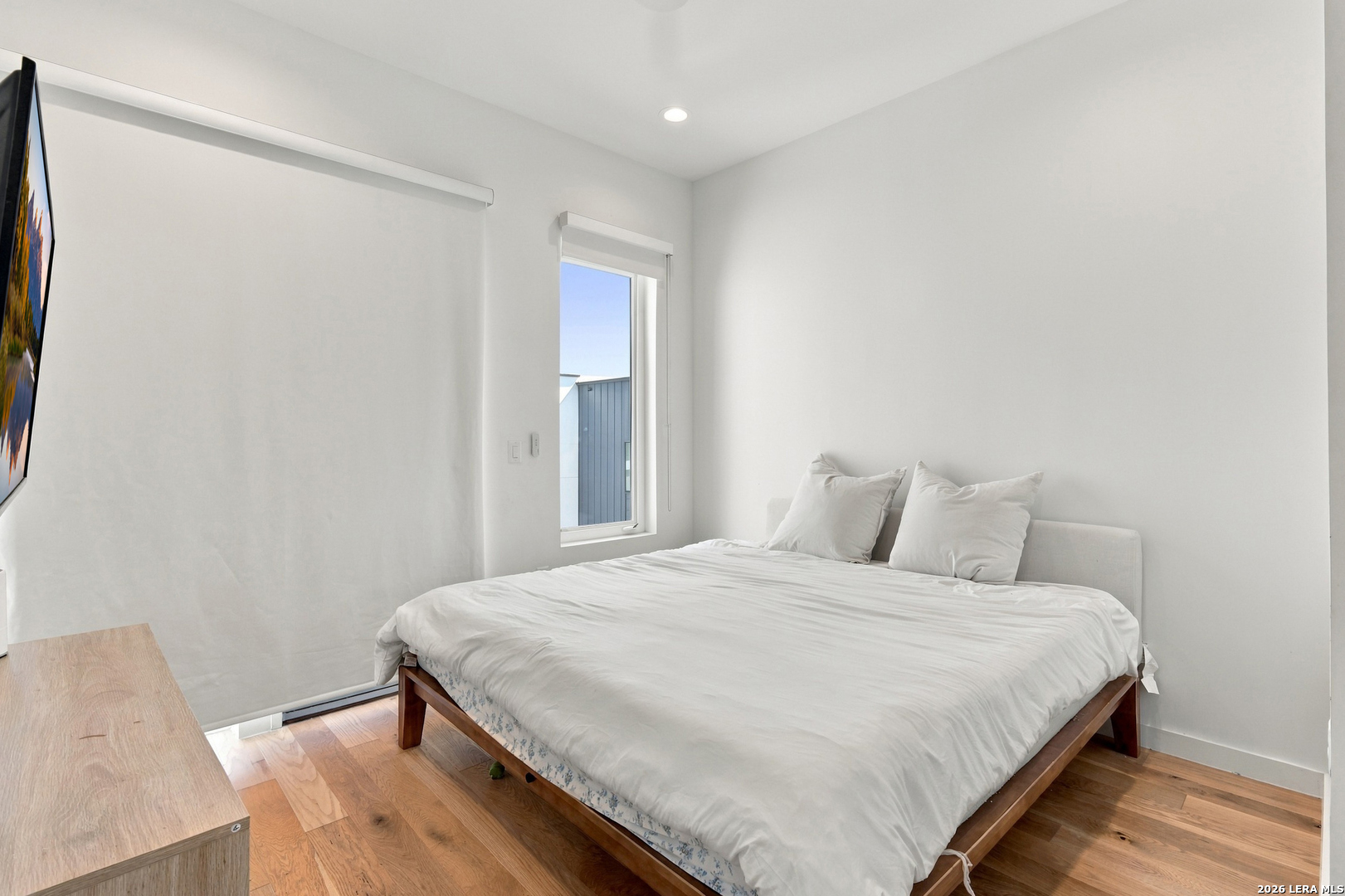 Furnished bedroom with a white bed, warm wood flooring, and a large roller shade.