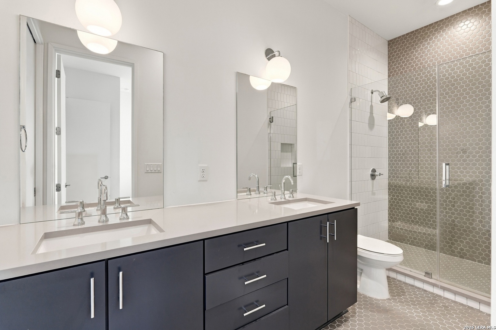 Modern bathroom with a double vanity and glass shower lined with gray hexagonal tile.