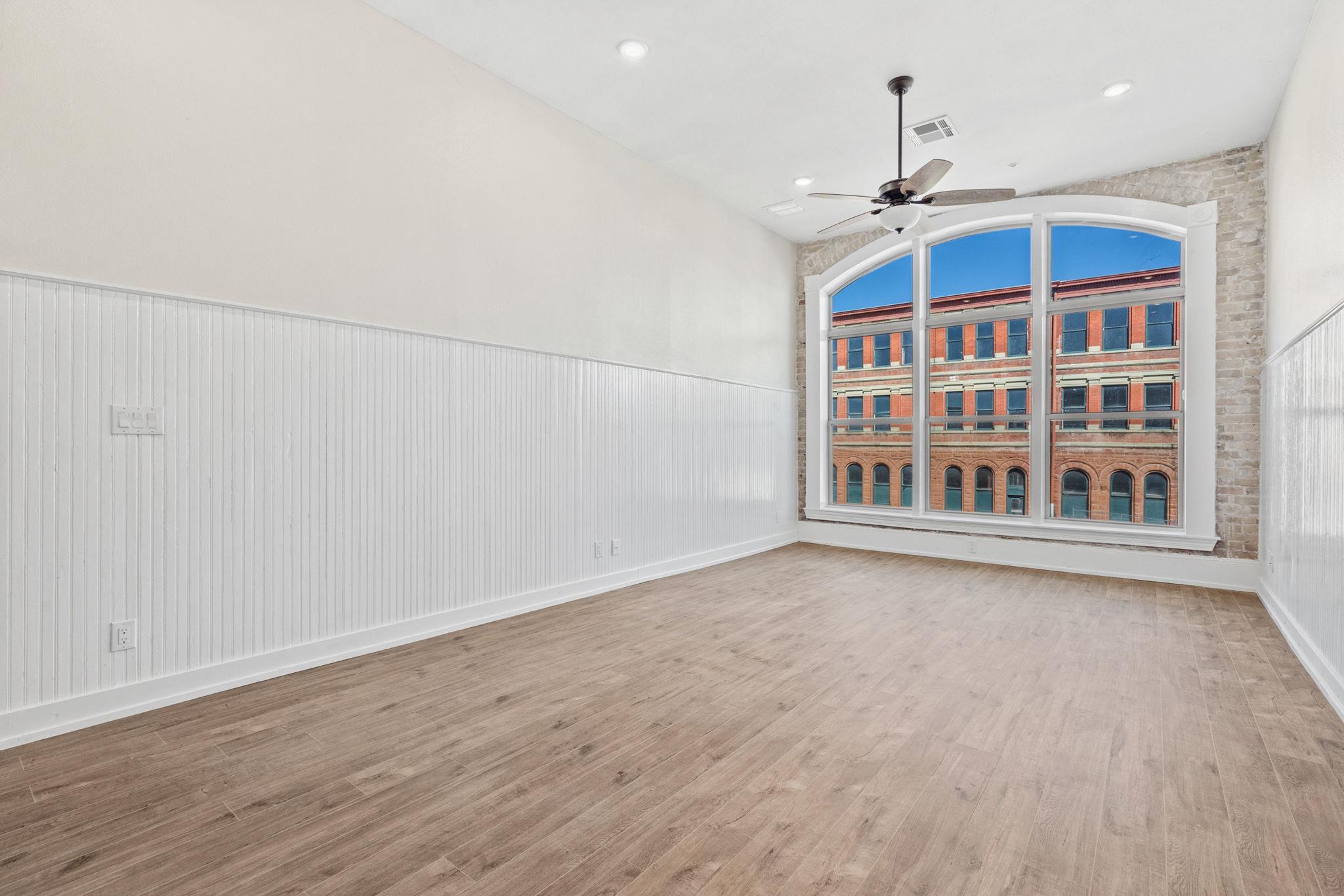Empty room with large arched windows, wood-look floors, white beadboard walls, and a ceiling fan.