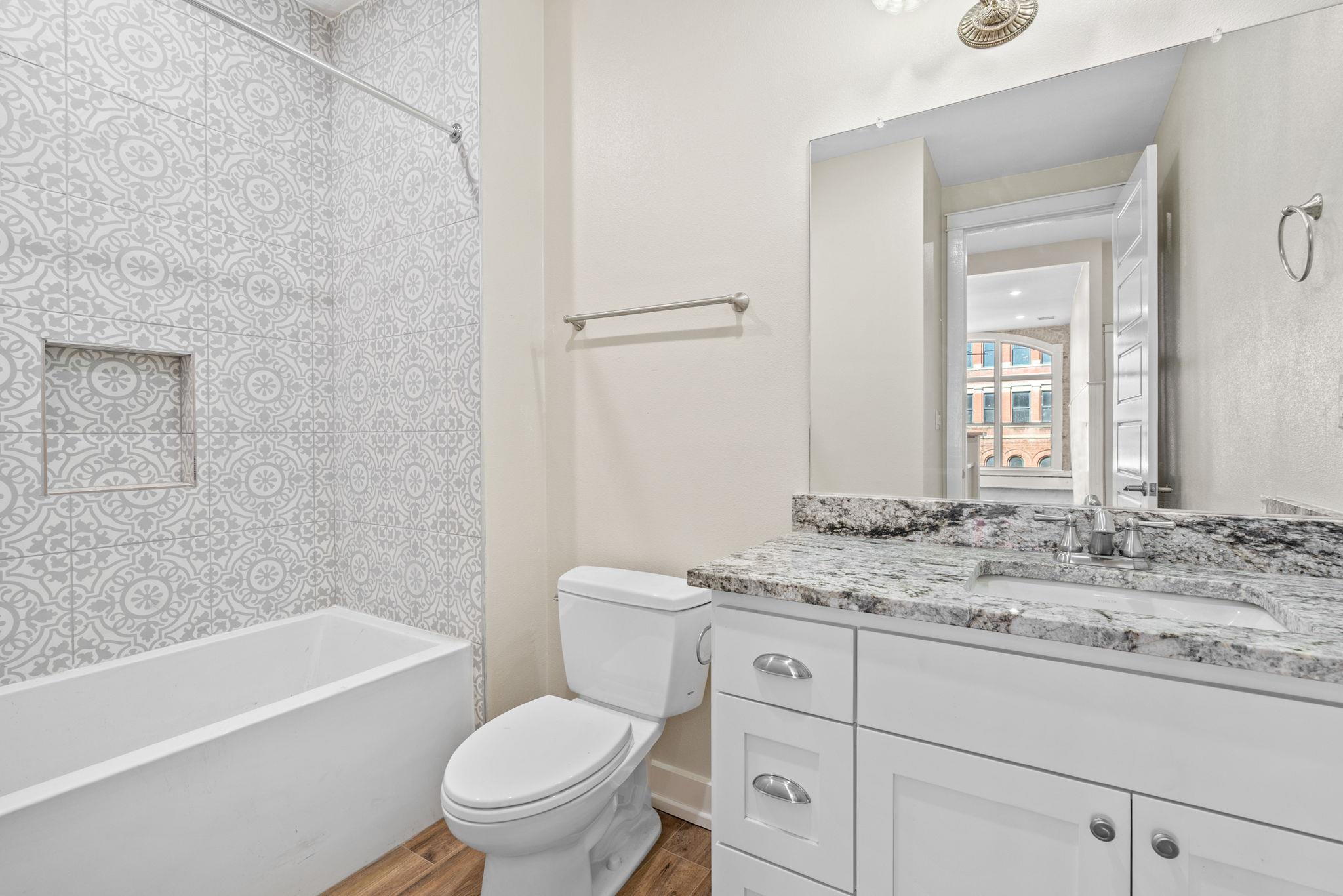 Bathroom with patterned tub surround, white vanity, granite countertop, and large mirror.