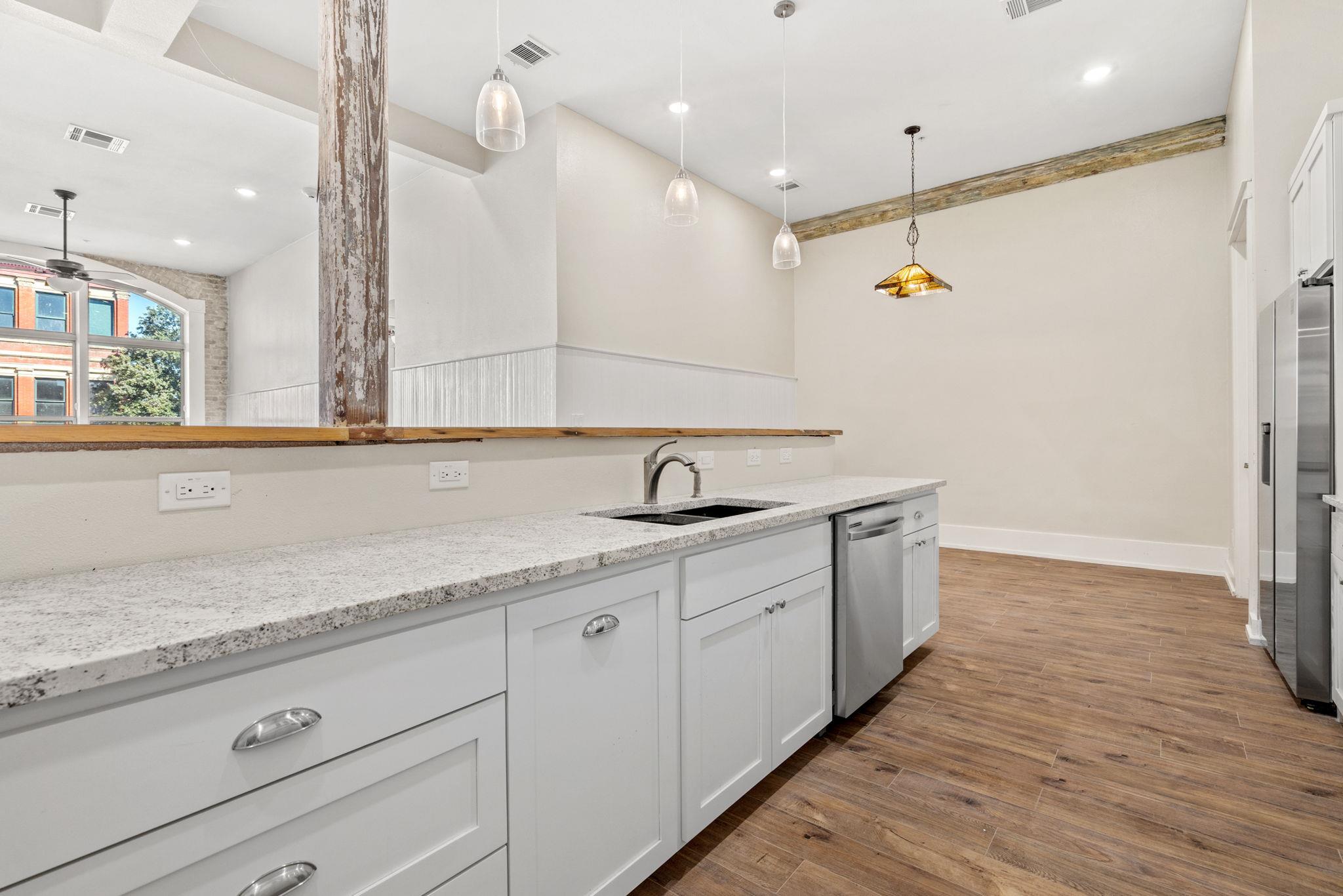 Kitchen with white cabinets and stone counters opening to a wood-floored dining area with pendant lights.