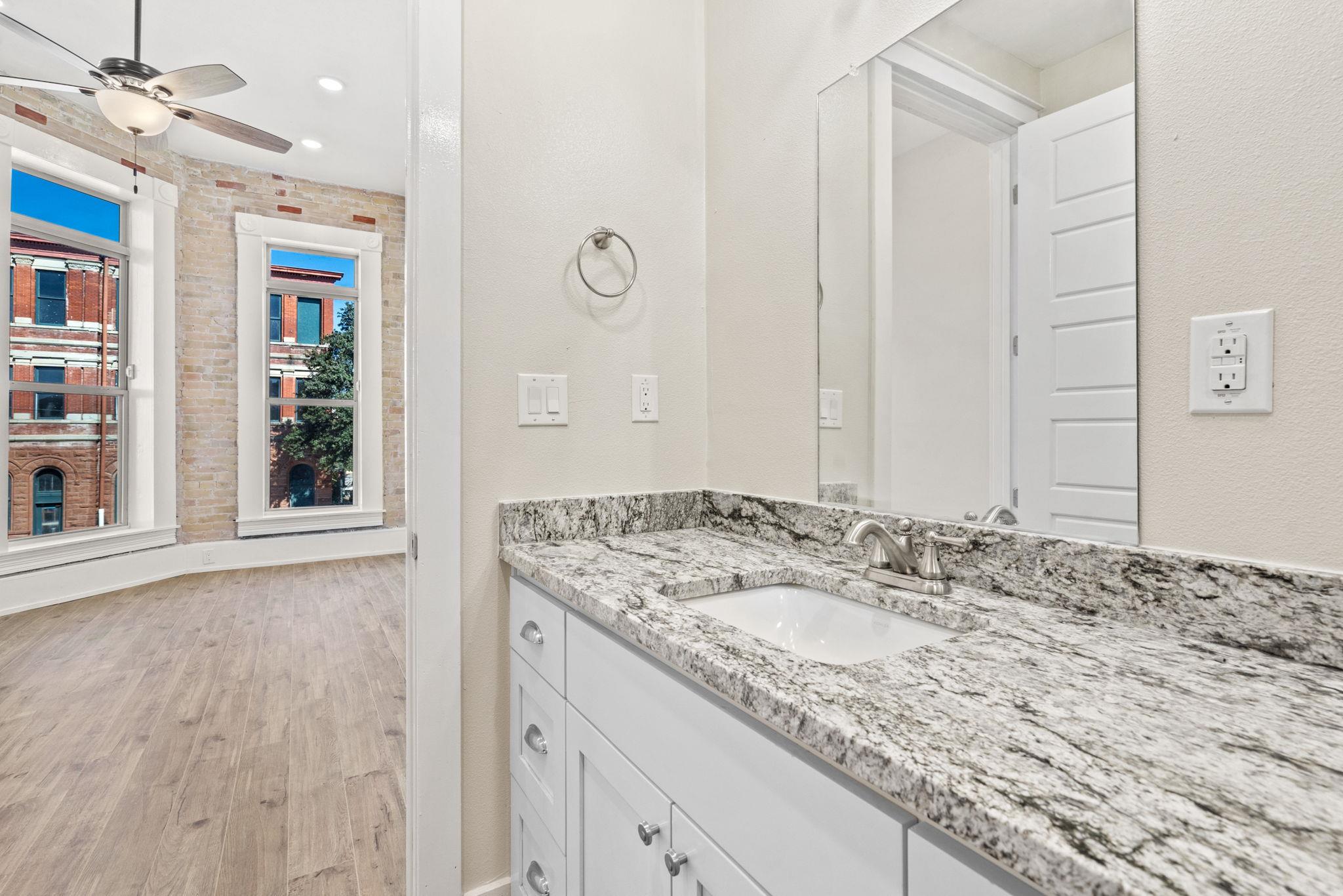 Bathroom vanity with granite countertop beside a bright room with tall windows and exposed brick.