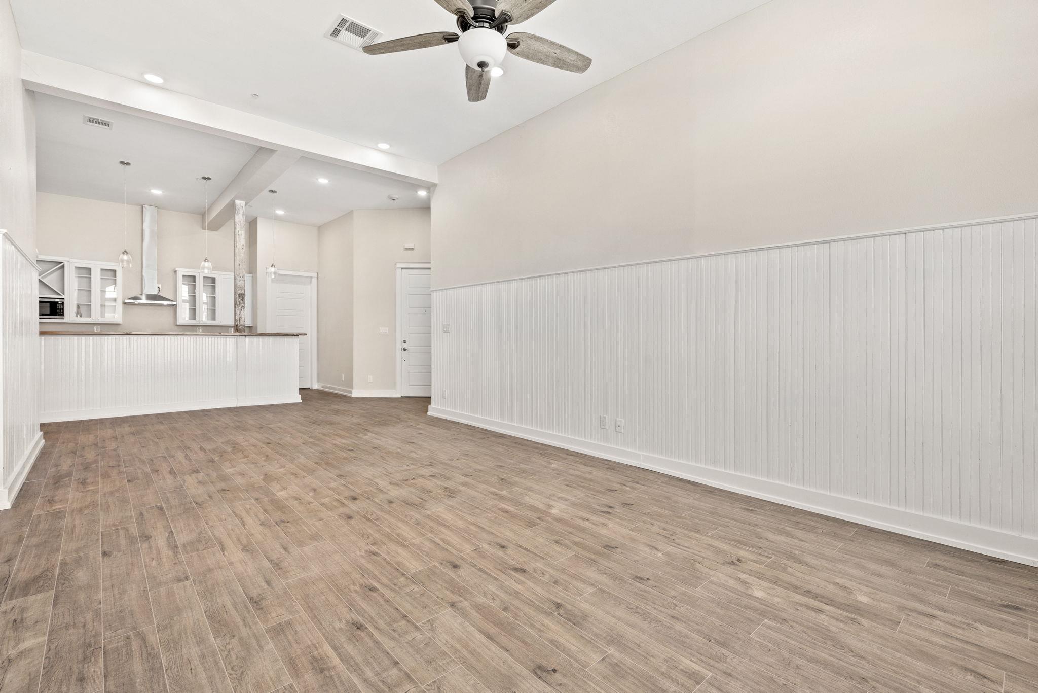 Empty open-plan living area with wood-look floors, white paneled walls, and a kitchen breakfast bar.
