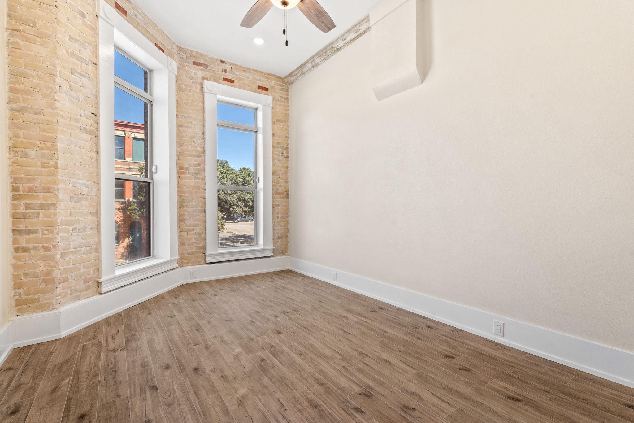 Empty room with exposed brick walls, tall windows, wood-look flooring, and a ceiling fan.