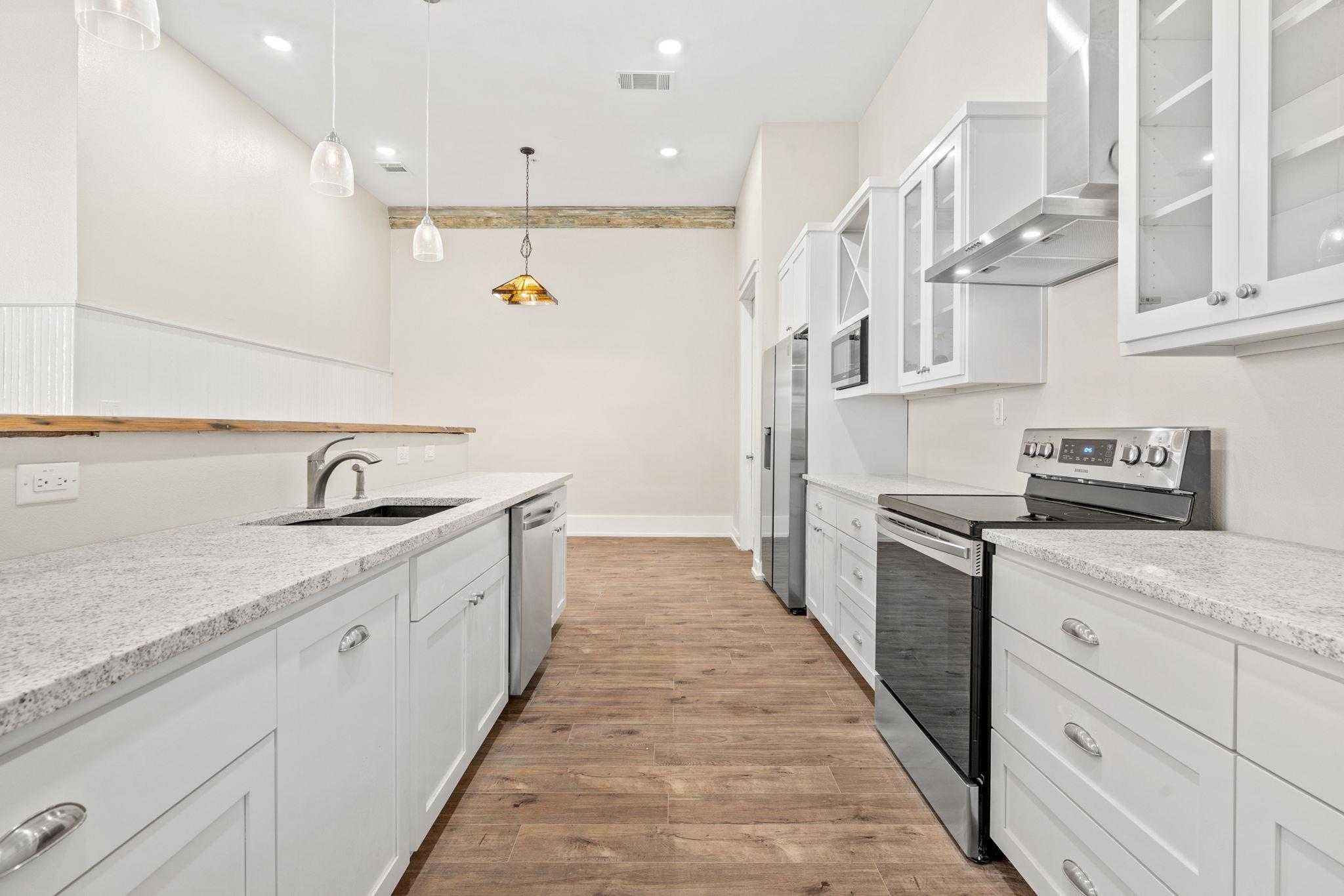 Vacant kitchen with white cabinetry, stainless steel appliances, stone counters, and warm wood-look flooring.