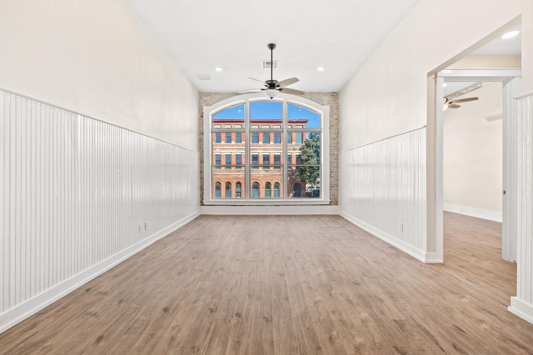 Empty room with wood-look floors, white beadboard walls, and a large arched window overlooking a brick building.