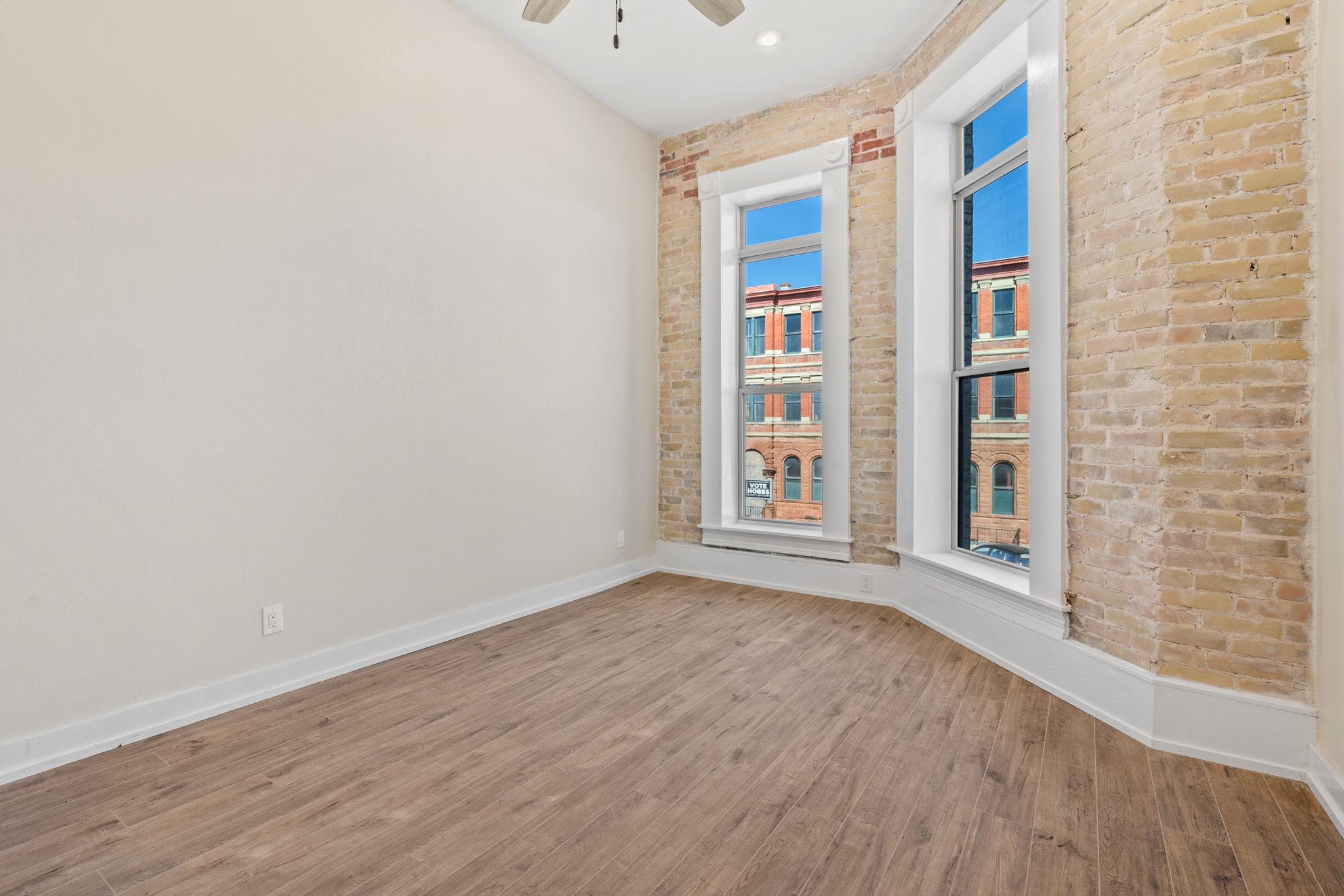 Vacant room with exposed brick, wood-look flooring, and two tall windows overlooking a brick building.