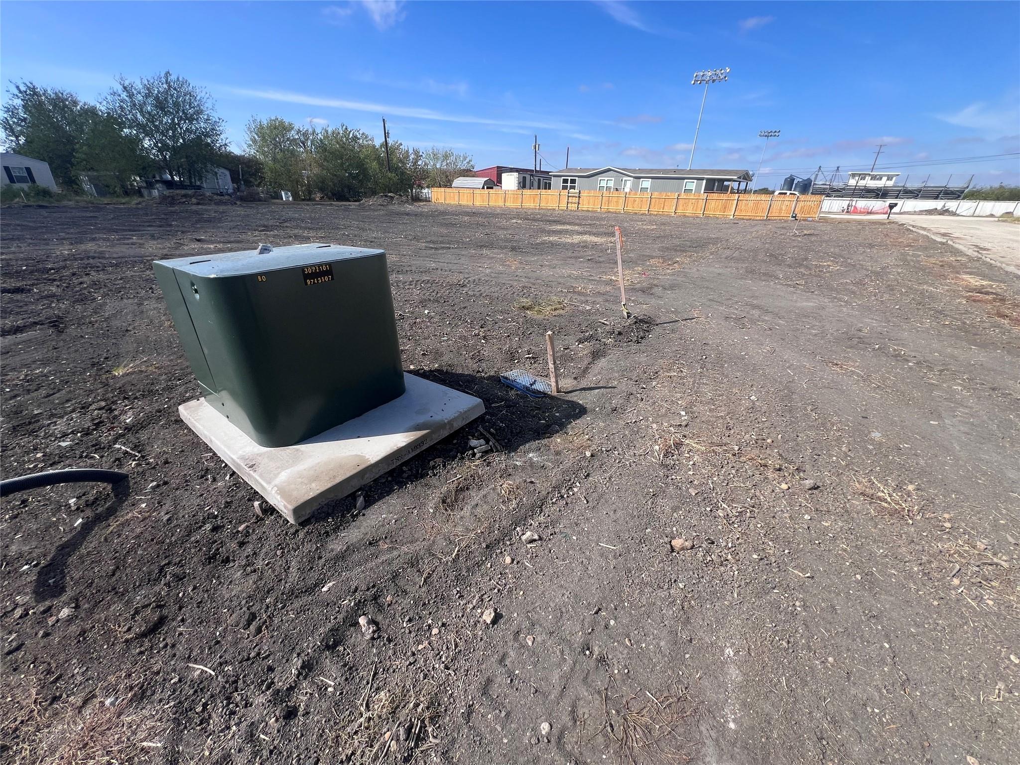 Cleared dirt lot with a green utility cabinet, survey stakes, and wooden fence.