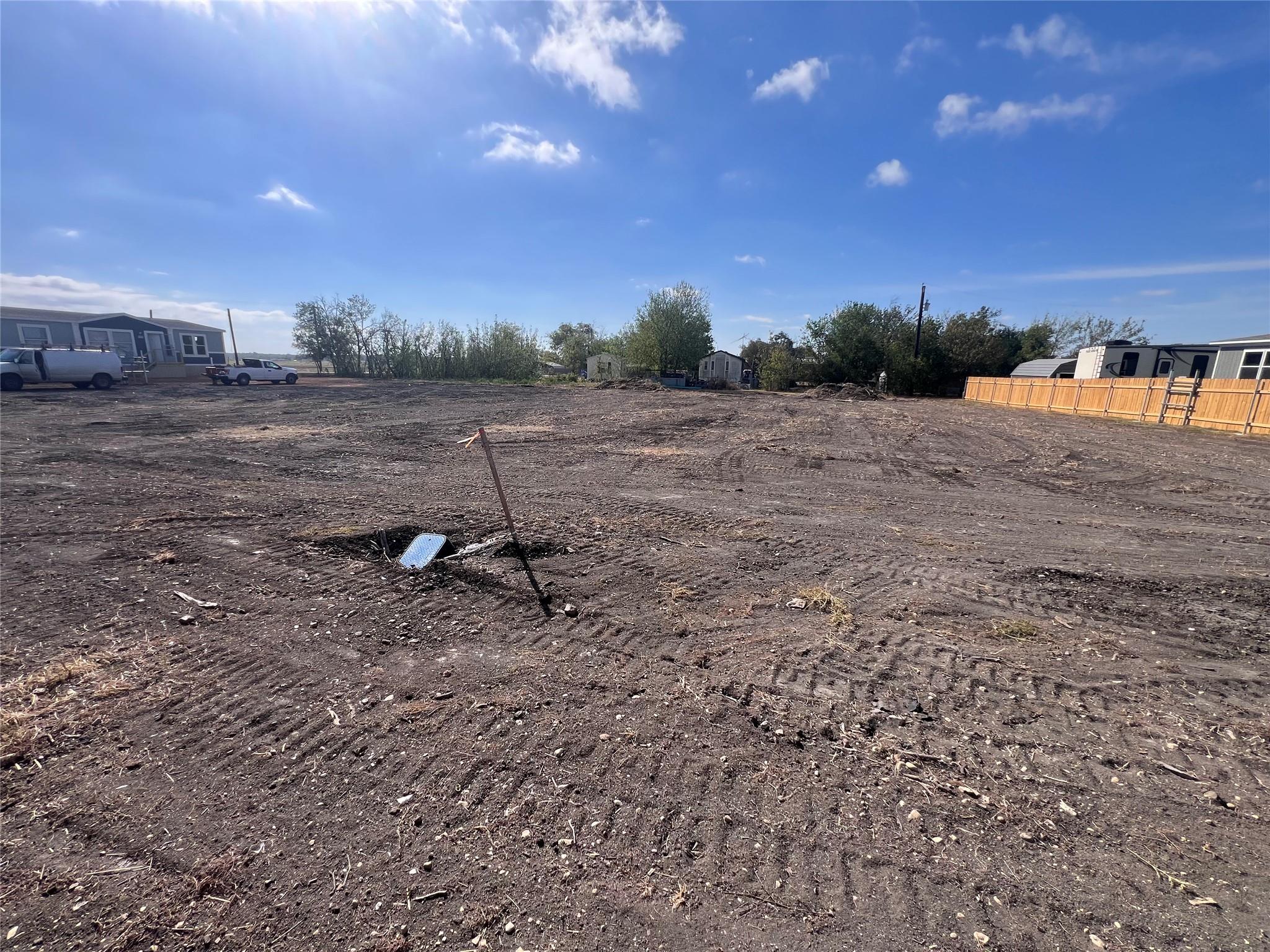 Cleared dirt lot with a utility marker, neighboring manufactured homes, and a wooden fence.