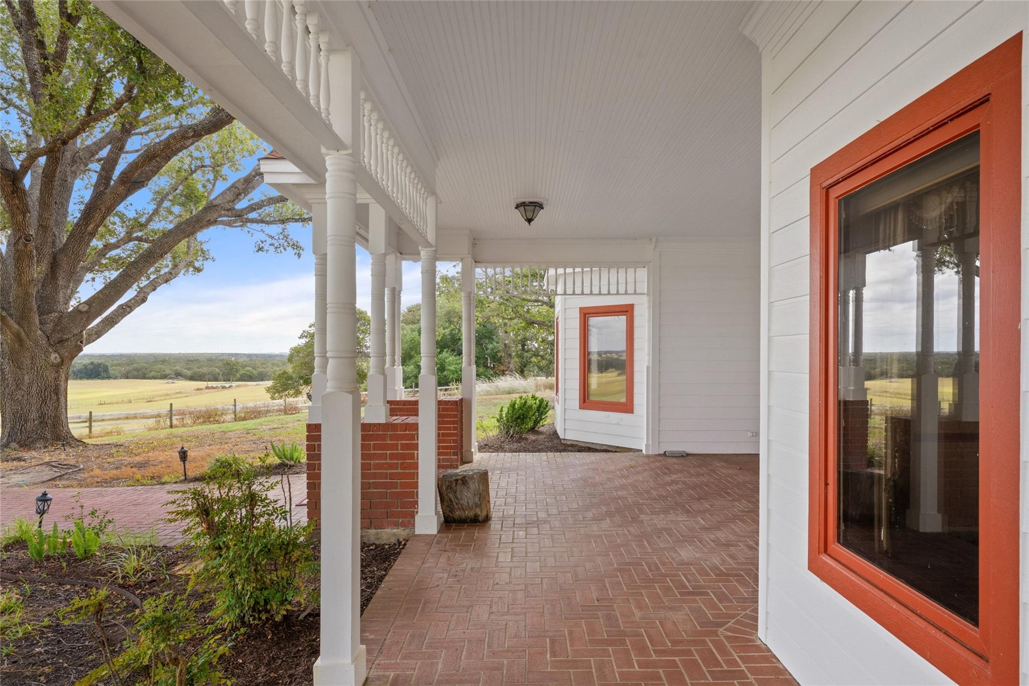 Farmhouse porch with white columns, brick pavers, red-trimmed windows, and pasture views.