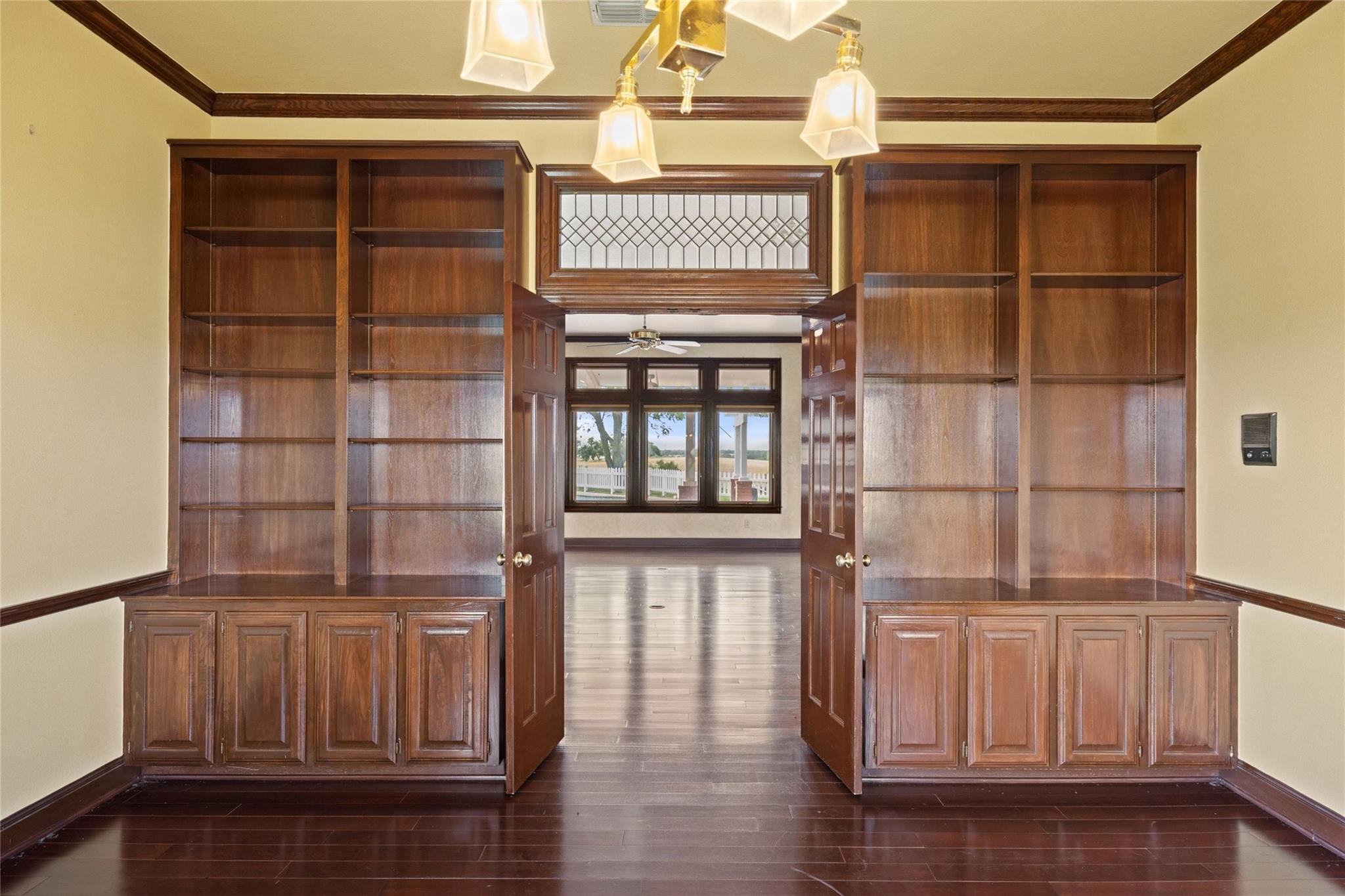 Unfurnished study with built-in dark wood bookshelves and open double doors to a bright room.