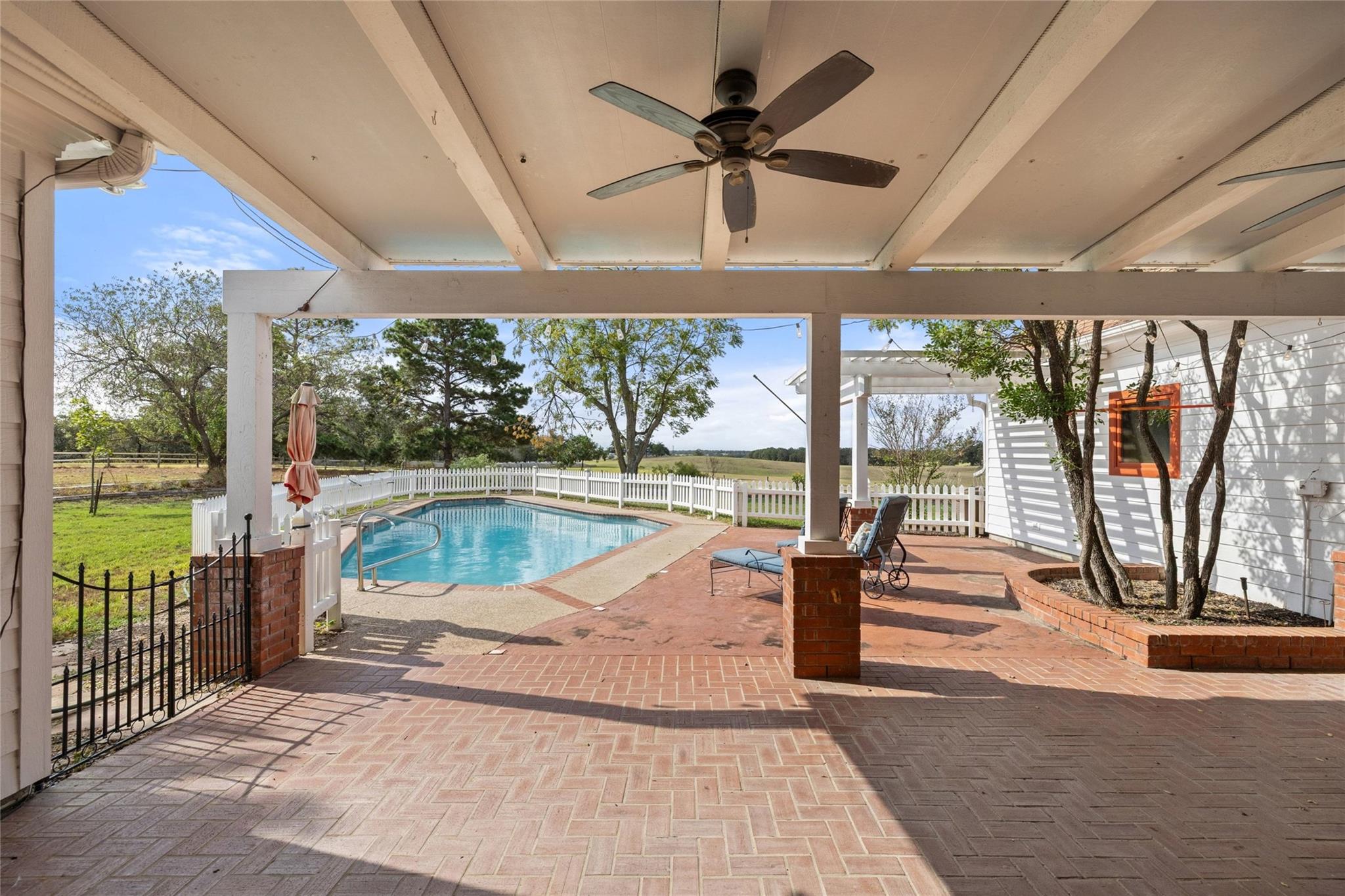 Swimming pool beside a covered brick patio with lounge chairs and white picket fencing.