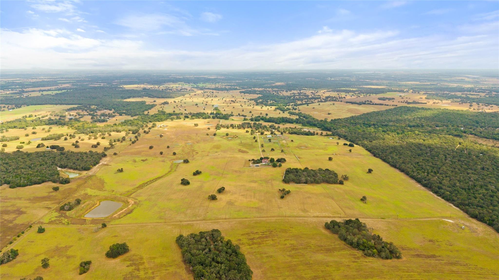 Aerial view of open pastureland with wooded areas, small ponds, and farm buildings.