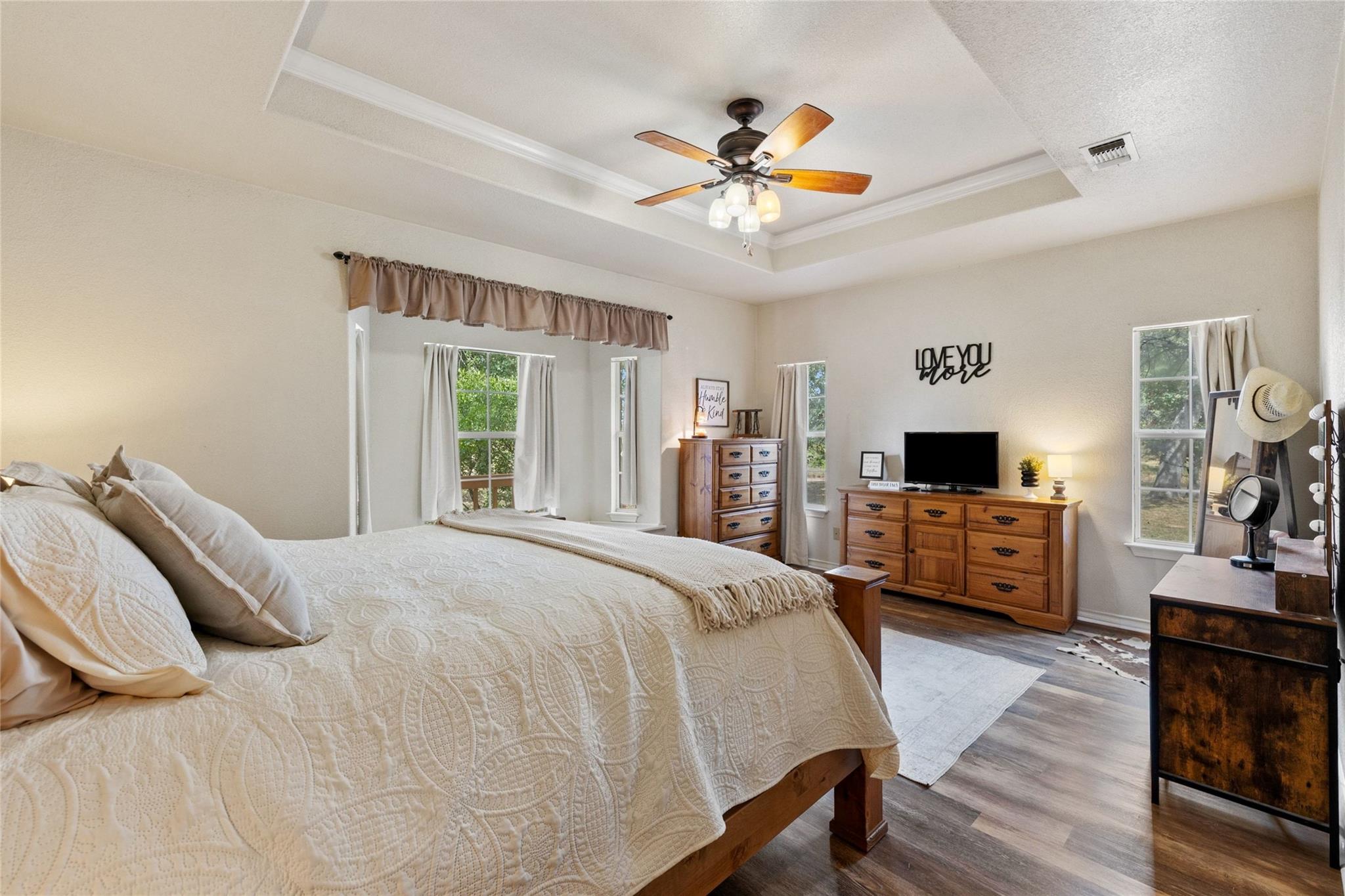 Furnished bedroom with a white quilted bed, tray ceiling, ceiling fan, and wooden dressers.