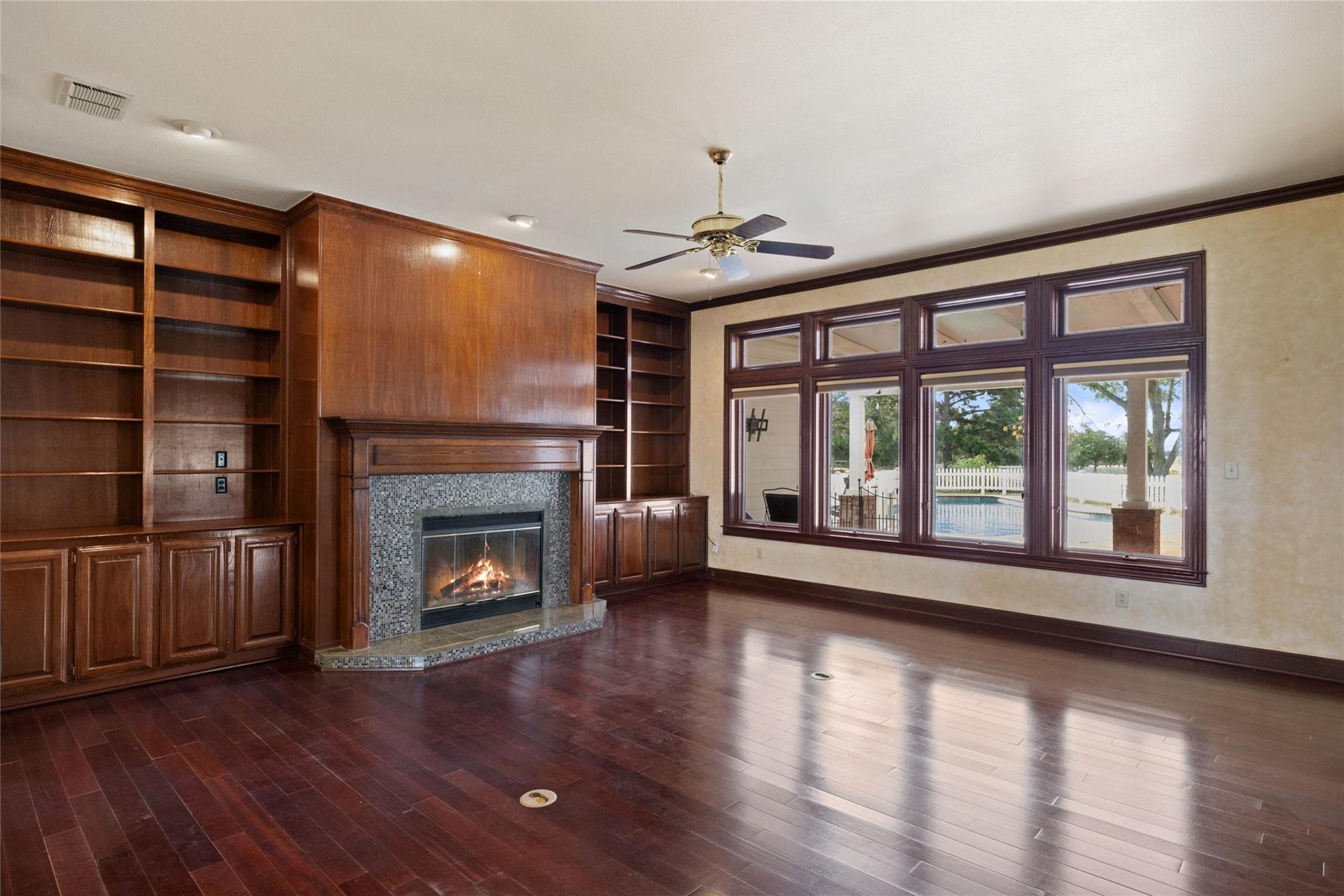Empty living room with built-in wood bookshelves, a lit fireplace, and large windows overlooking a pool.