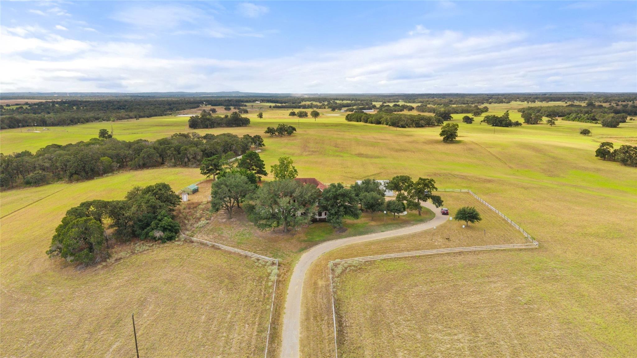 Aerial view of a tree-shaded rural home, fenced yard, long driveway, and surrounding open fields.