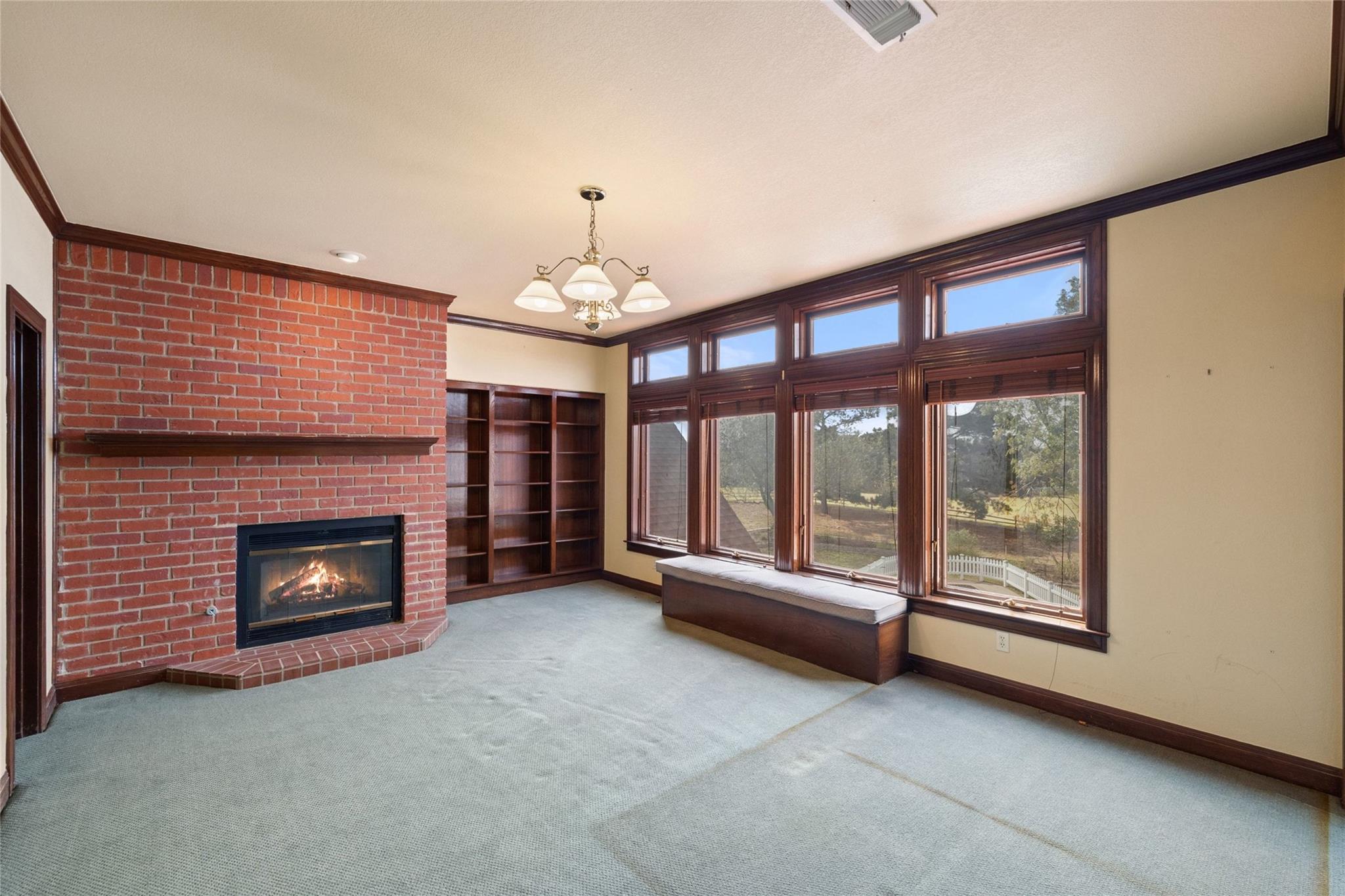 Empty living room with a brick fireplace, built-in bookshelves, and large windows overlooking trees.