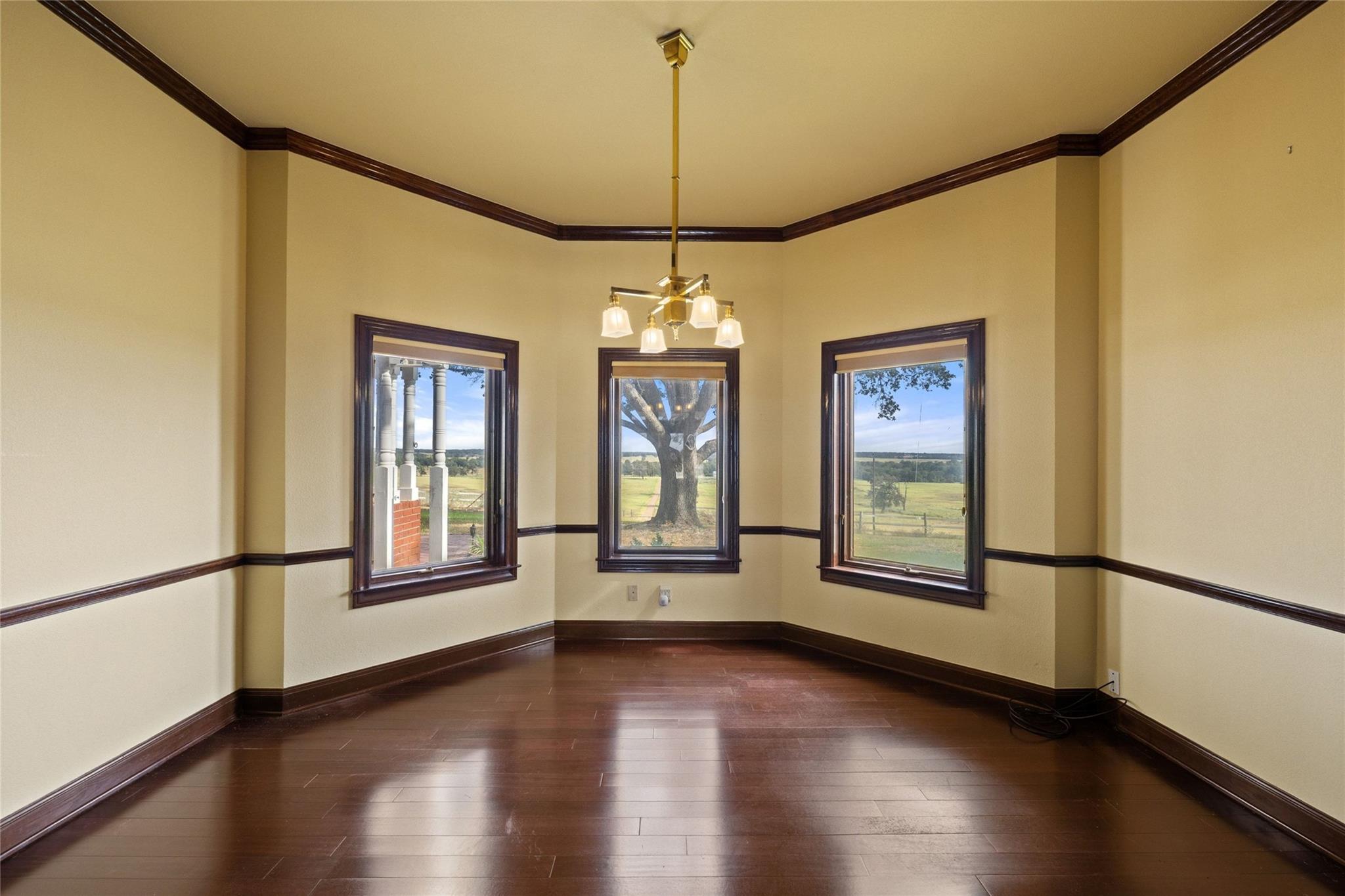Empty room with dark wood floors, a chandelier, and three windows overlooking open fields.