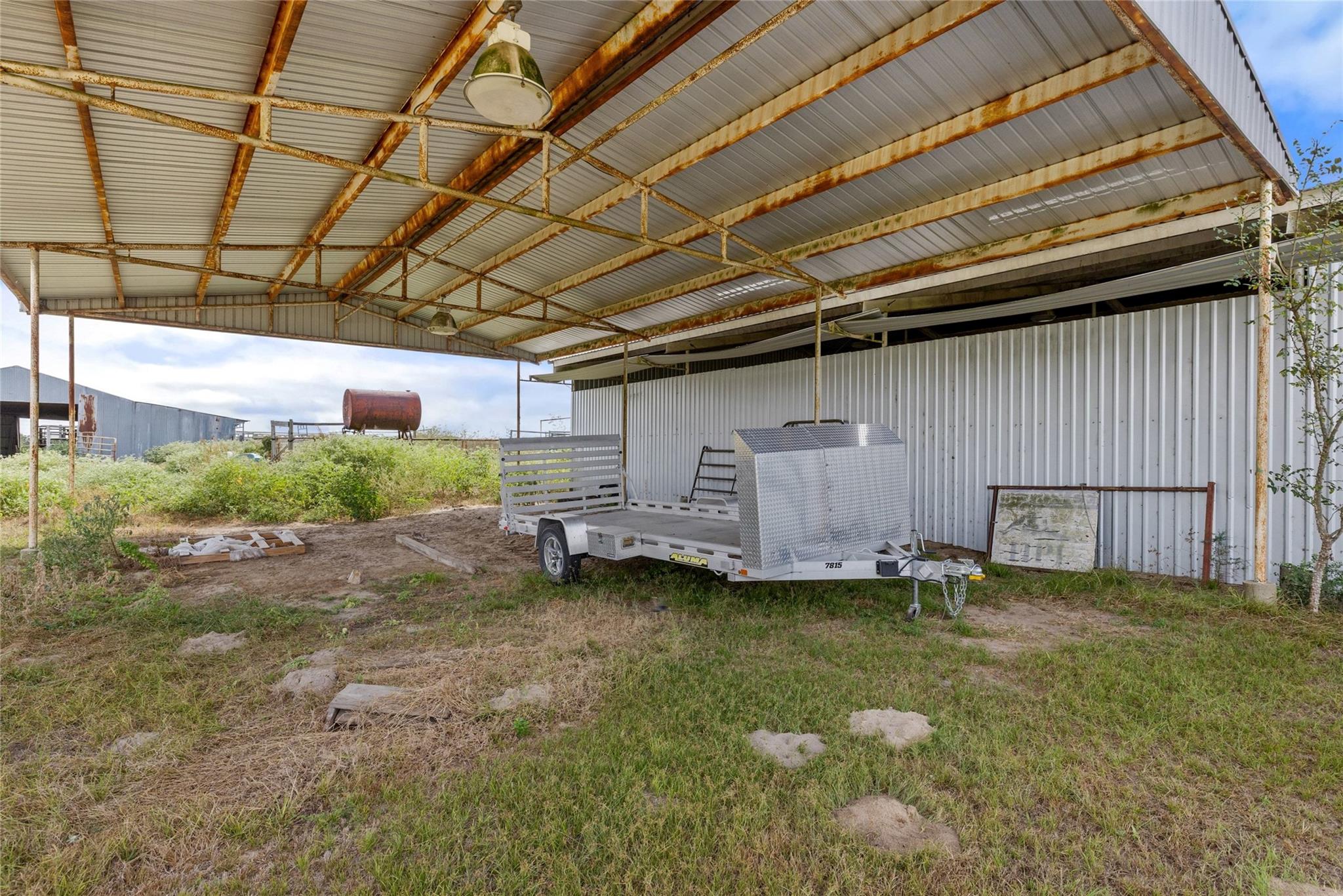 Open-sided metal shelter with a utility trailer beside a corrugated metal outbuilding.