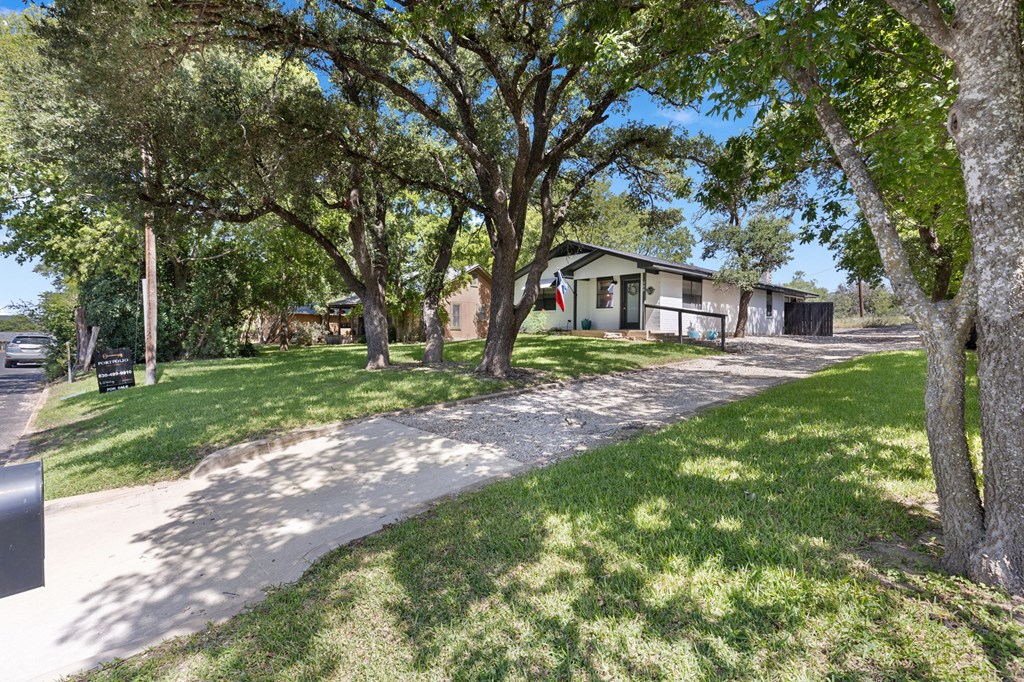 Single-story house behind a gravel driveway, shaded by mature trees, with a Texas flag by the porch.