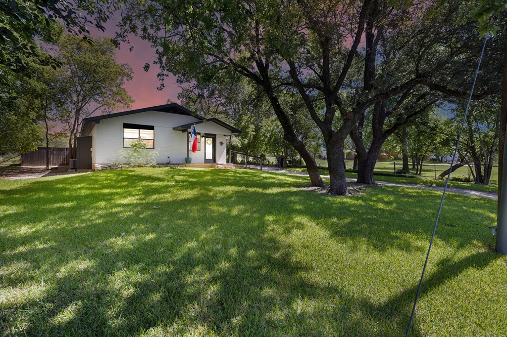 Single-story white house beneath mature oak trees, with a Texas flag by the entrance and a broad grassy lawn.