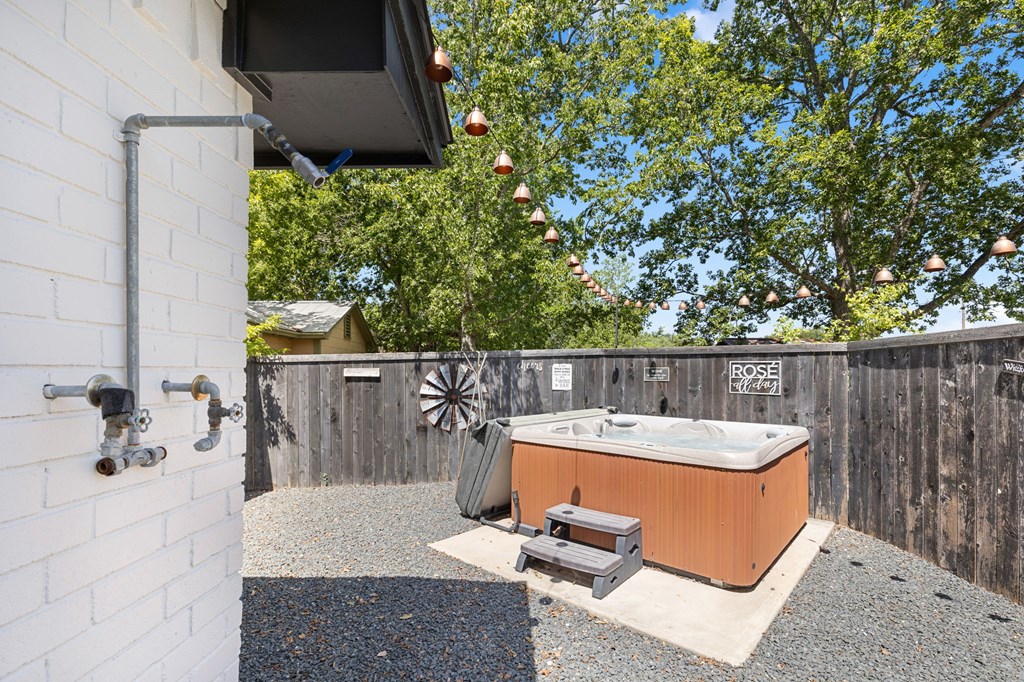 Outdoor hot tub on a gravel patio, enclosed by a wooden fence beneath leafy trees and string lights.