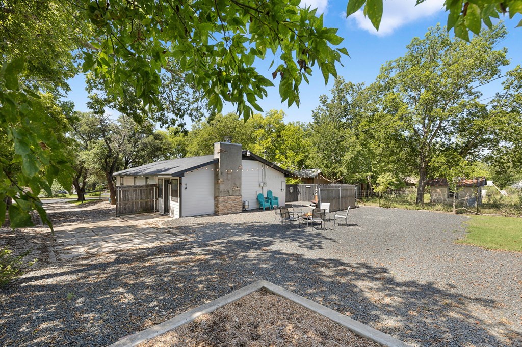 White house with a stone chimney beside a shaded gravel yard and outdoor seating.