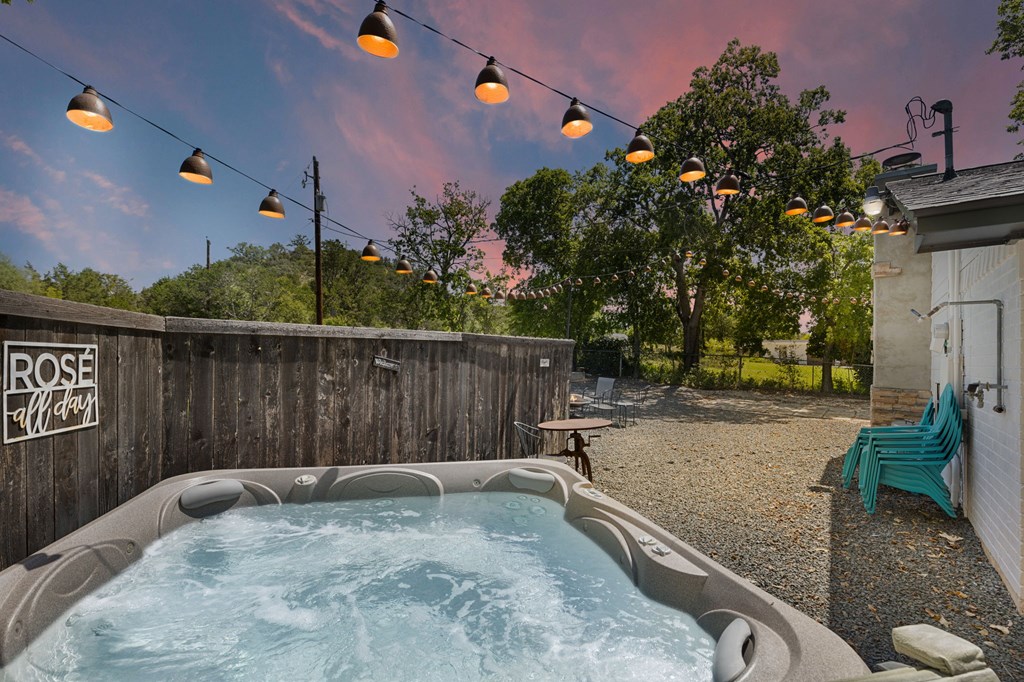 Outdoor hot tub on a gravel patio beneath string lights, with turquoise chairs and a pink dusk sky.