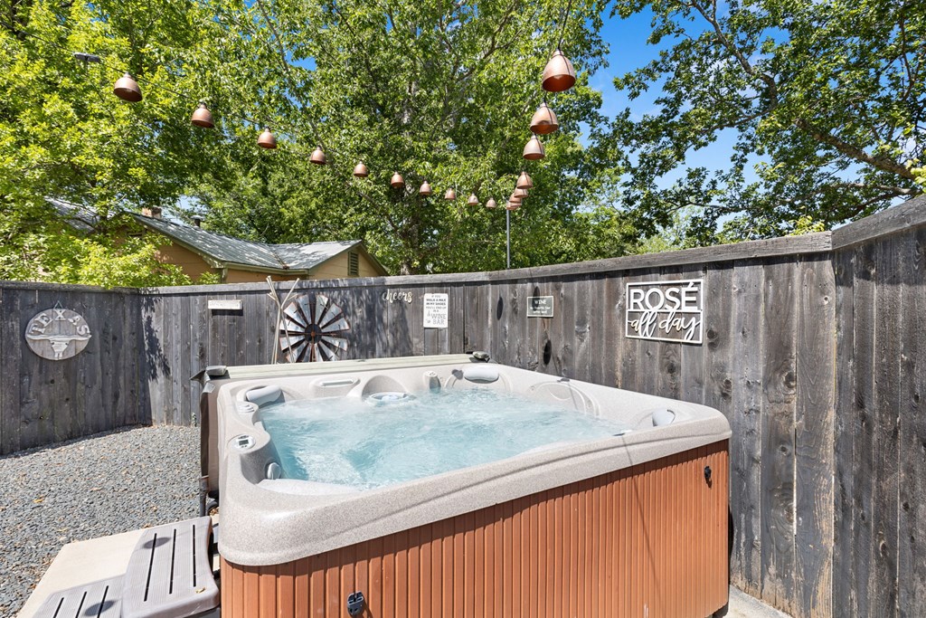 Outdoor hot tub in a fenced gravel patio beneath string lights and leafy trees.