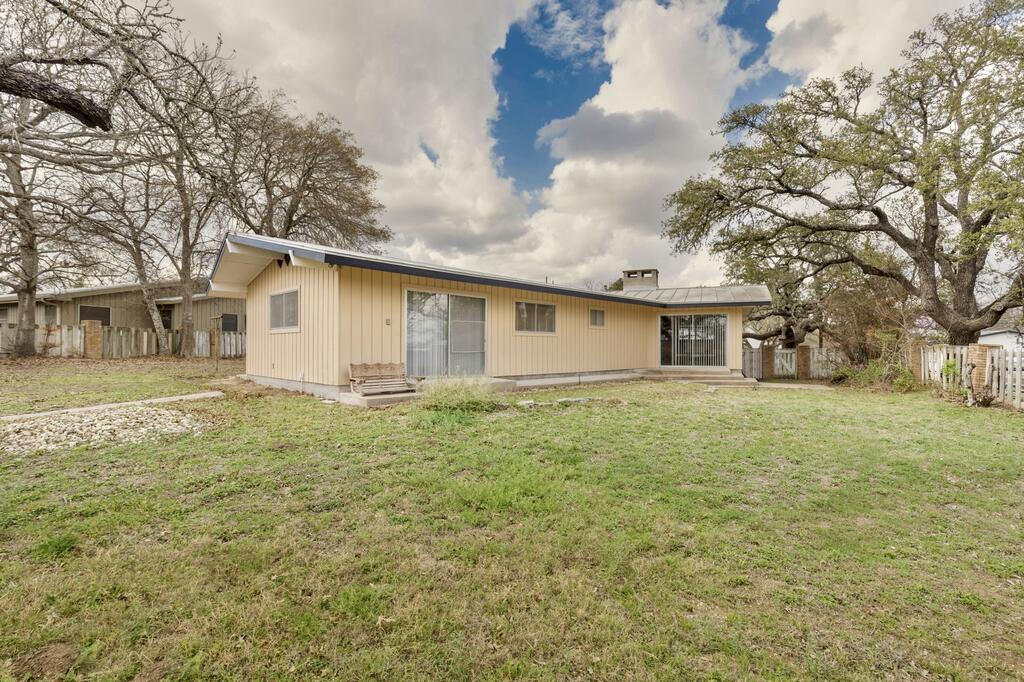Single-story beige house viewed from the backyard, with sliding doors and mature trees along the fence.