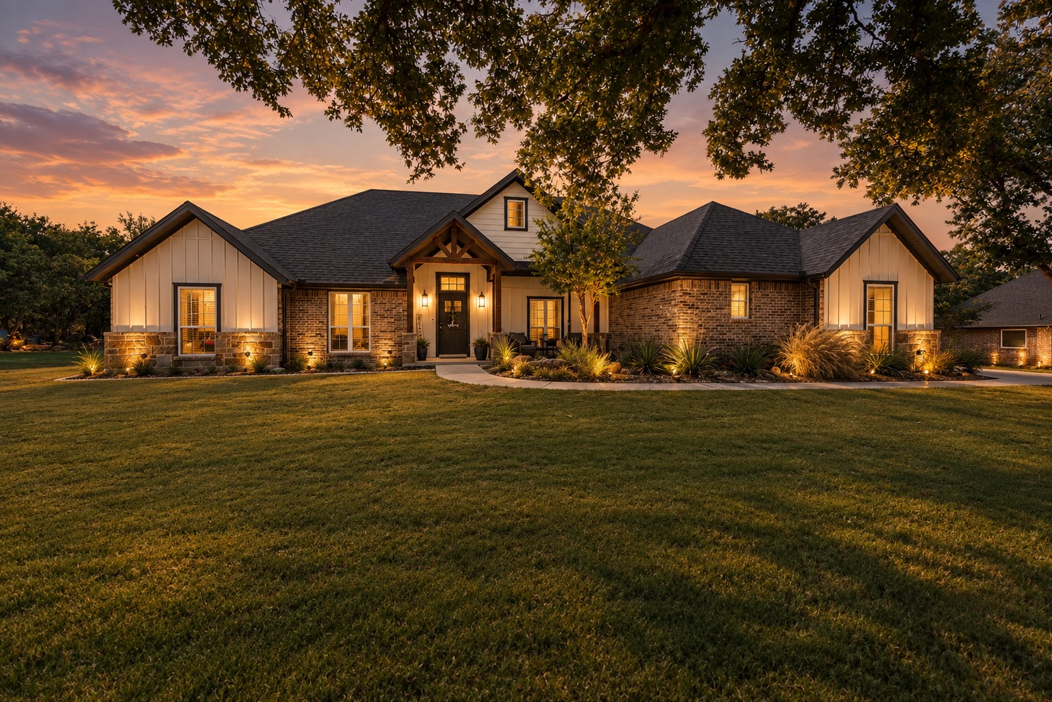 Single-story brick and siding home with glowing windows, landscaped beds, and a broad lawn at sunset.