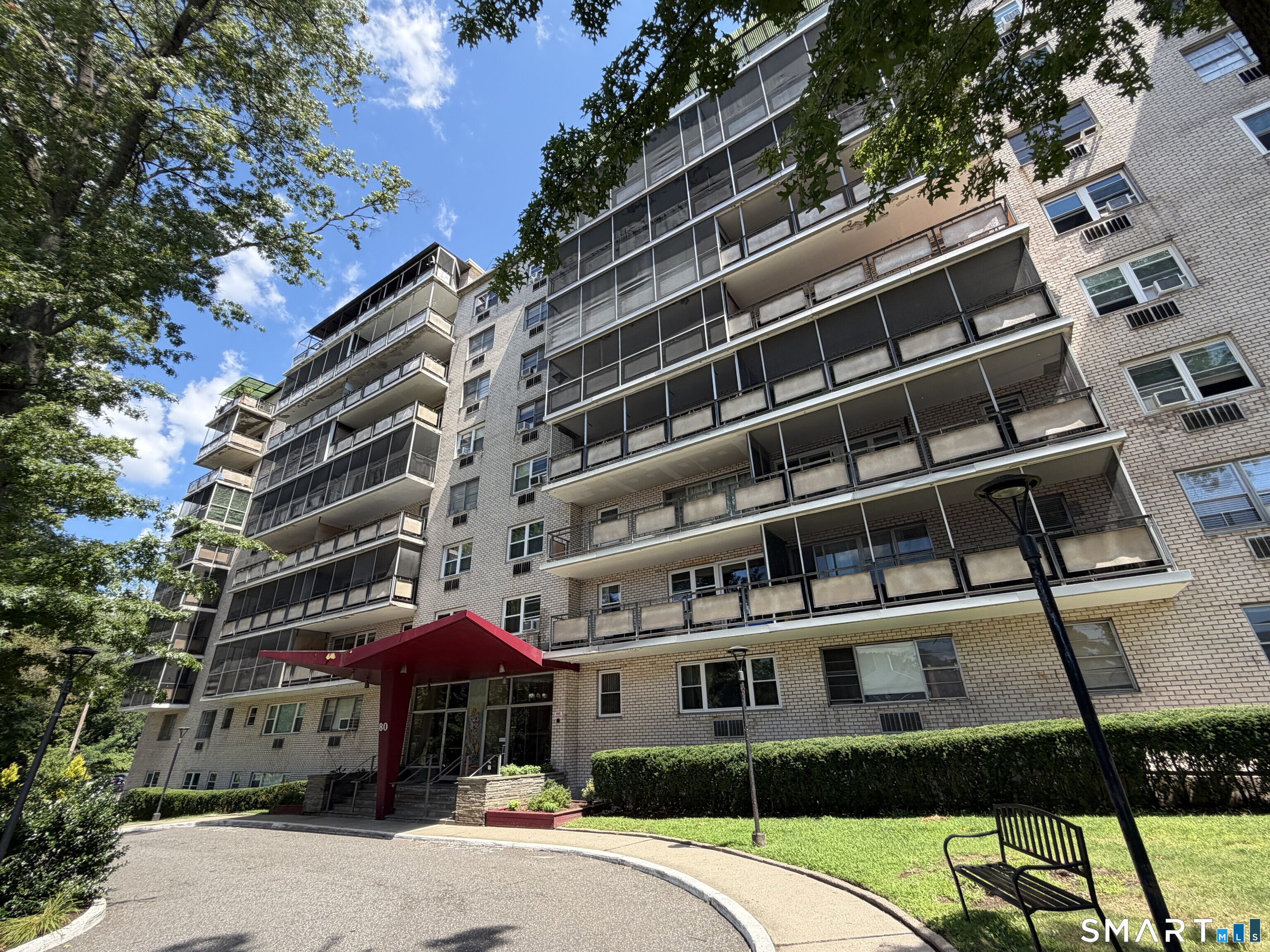 Brick apartment building with rows of balconies and a red entrance canopy, framed by leafy trees.