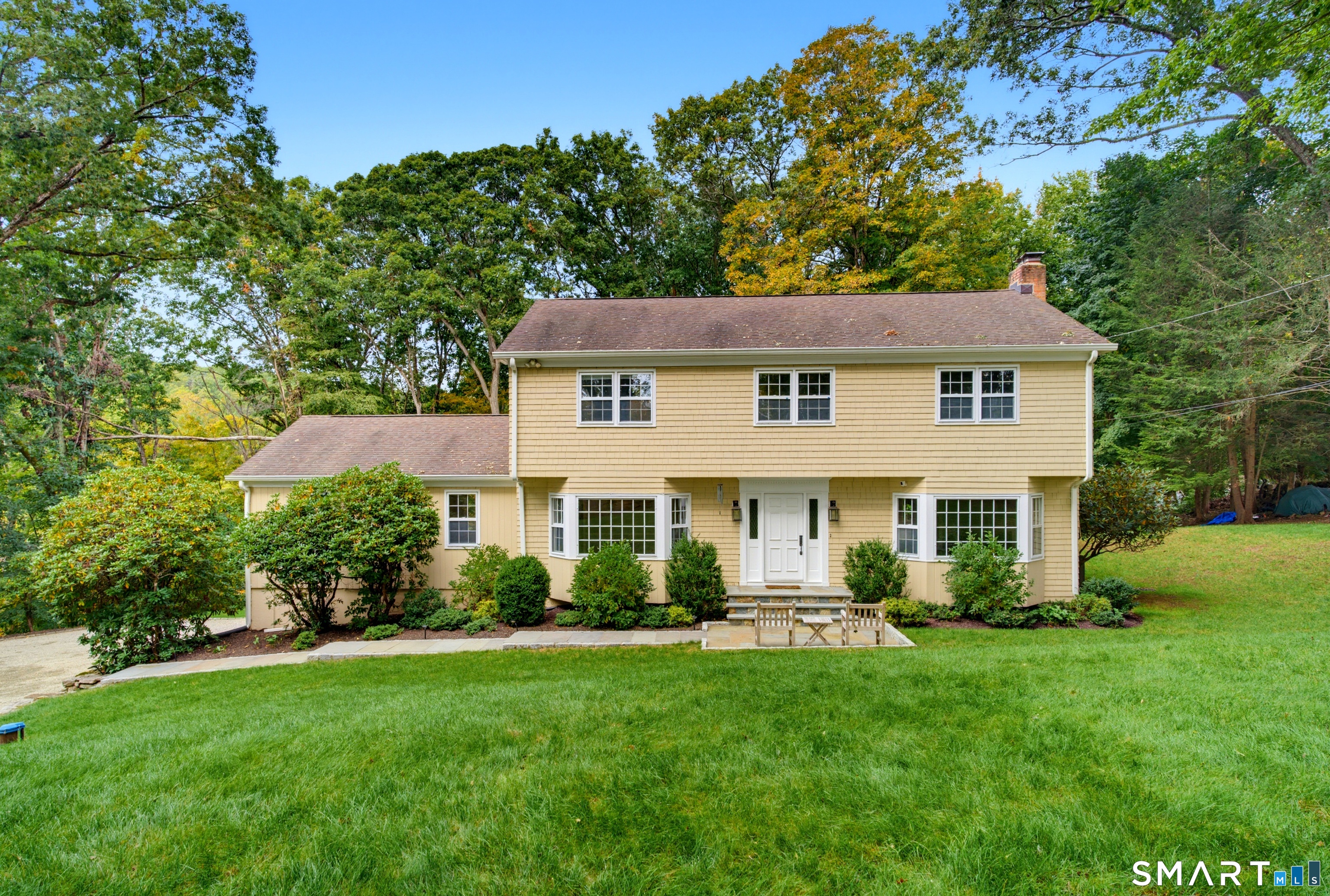 Two-story yellow-sided home with a small patio and broad lawn, surrounded by mature trees.
