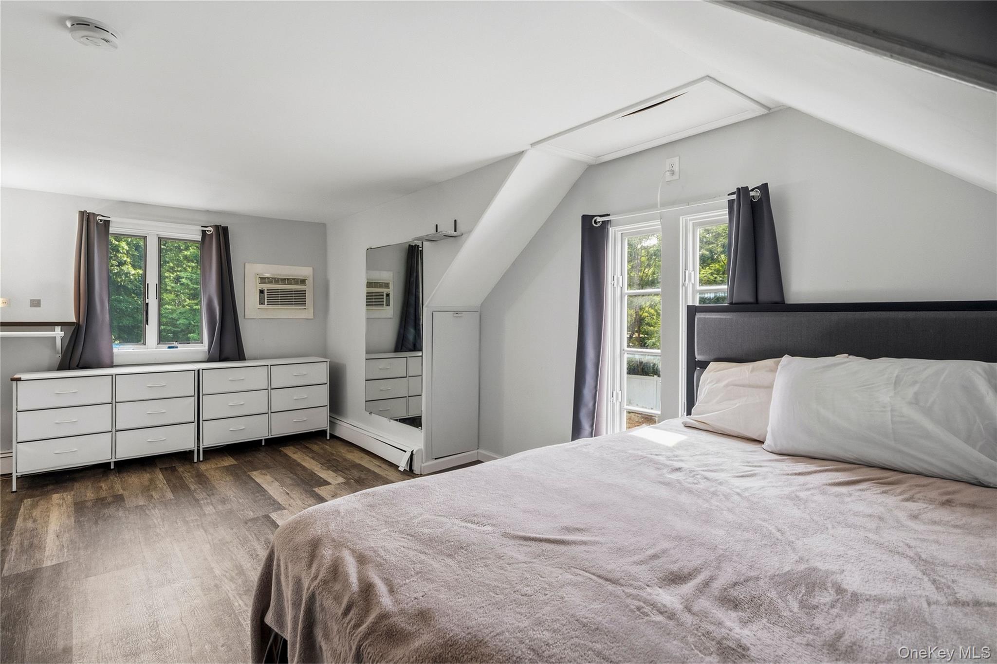 Bedroom with sloped ceiling, dark headboard, white dressers, and windows overlooking greenery.