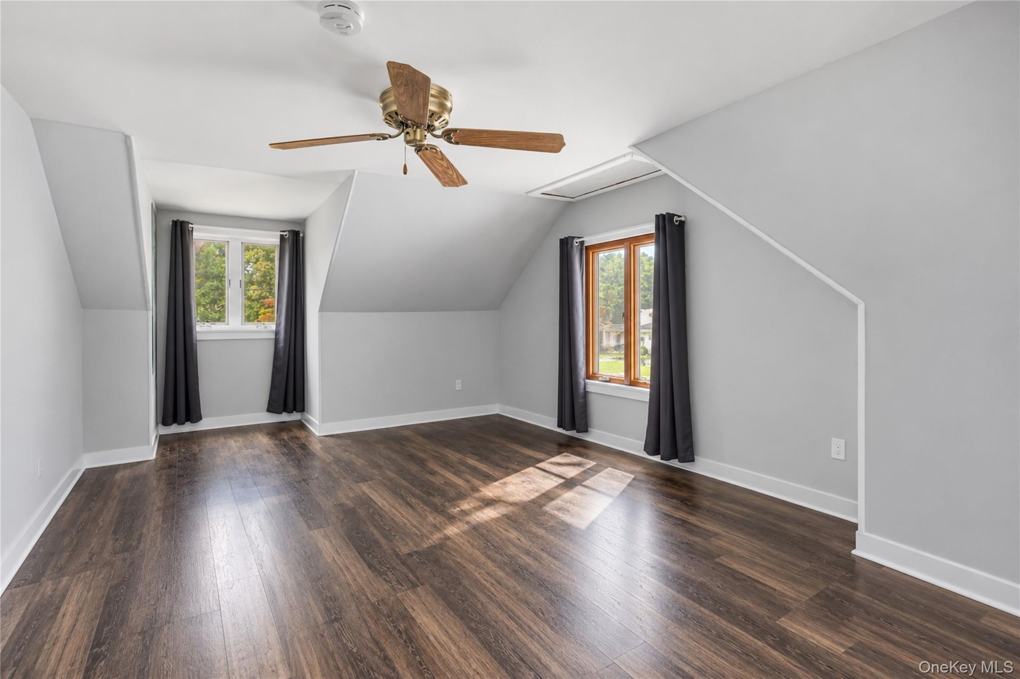 Vacant upper-level room with sloped ceilings, dark flooring, two curtained windows, and a ceiling fan.