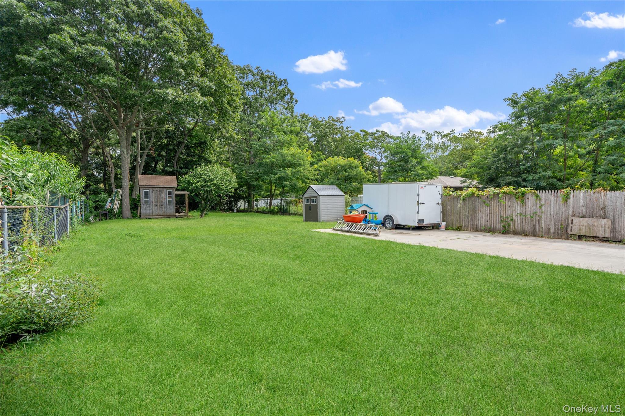 Backyard lawn bordered by mature trees, with two sheds and a white utility trailer.