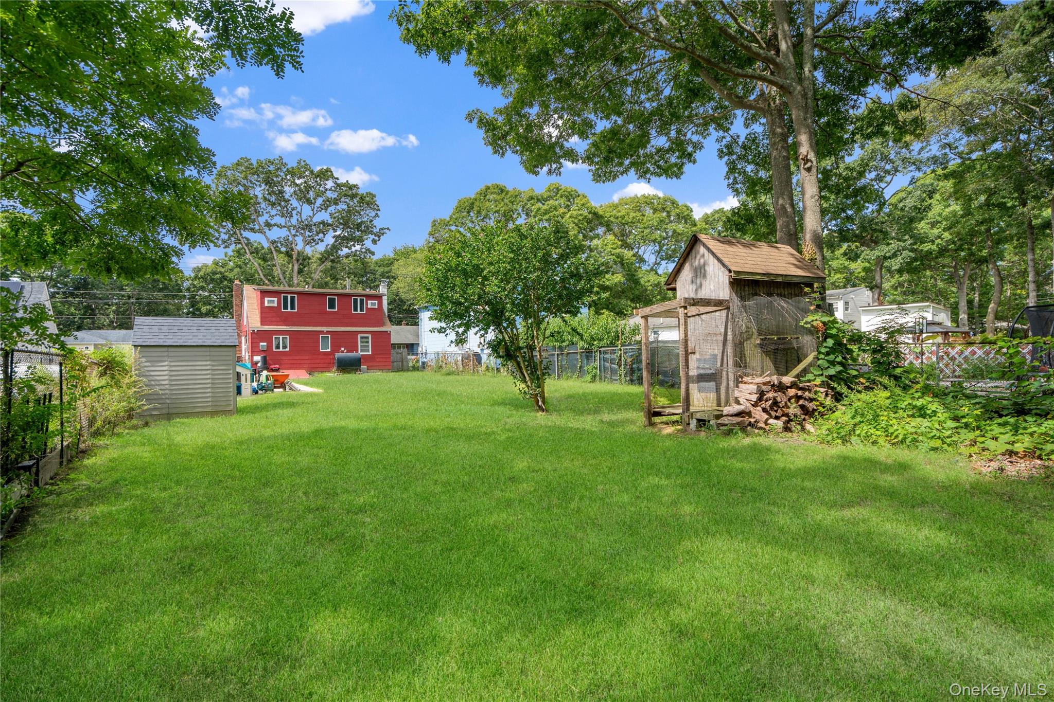 Spacious grassy backyard with mature trees, a shed, and a wooden outbuilding beside stacked firewood.