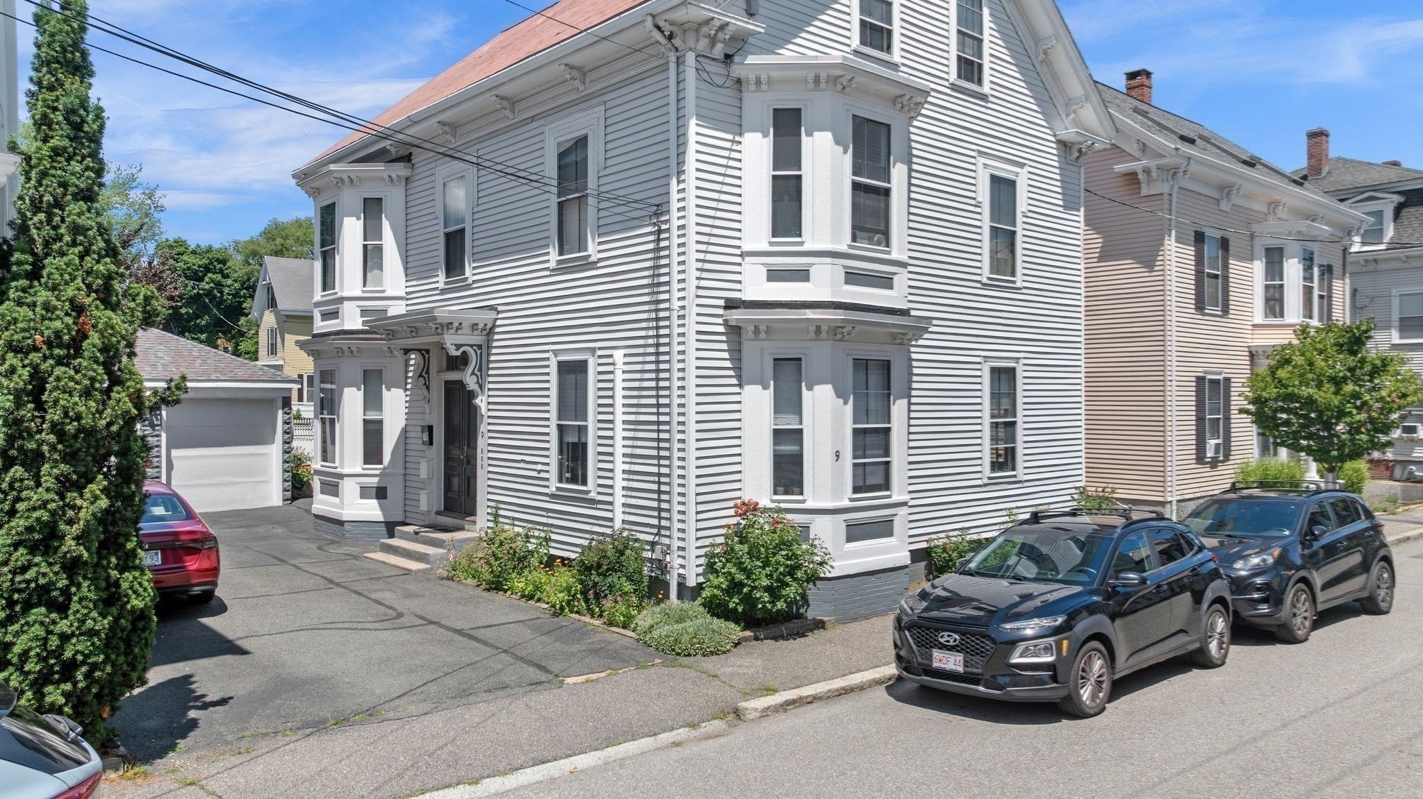 Gray clapboard multi-unit house with ornate white trim, bay windows, and cars parked along the street.