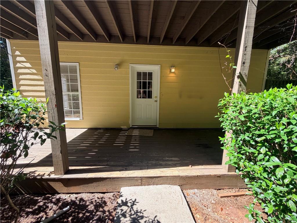 Covered wooden porch with a white entry door, yellow siding, and surrounding shrubs.