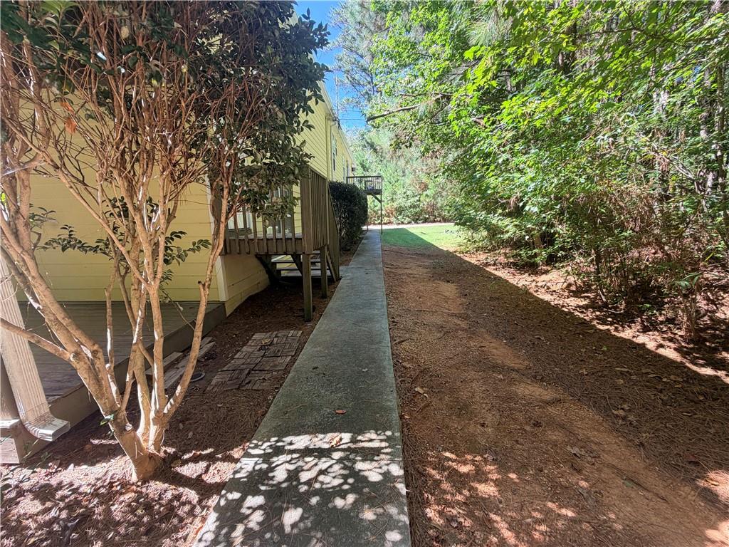 Pale yellow house beside a concrete walkway bordered by dense trees and shrubs.
