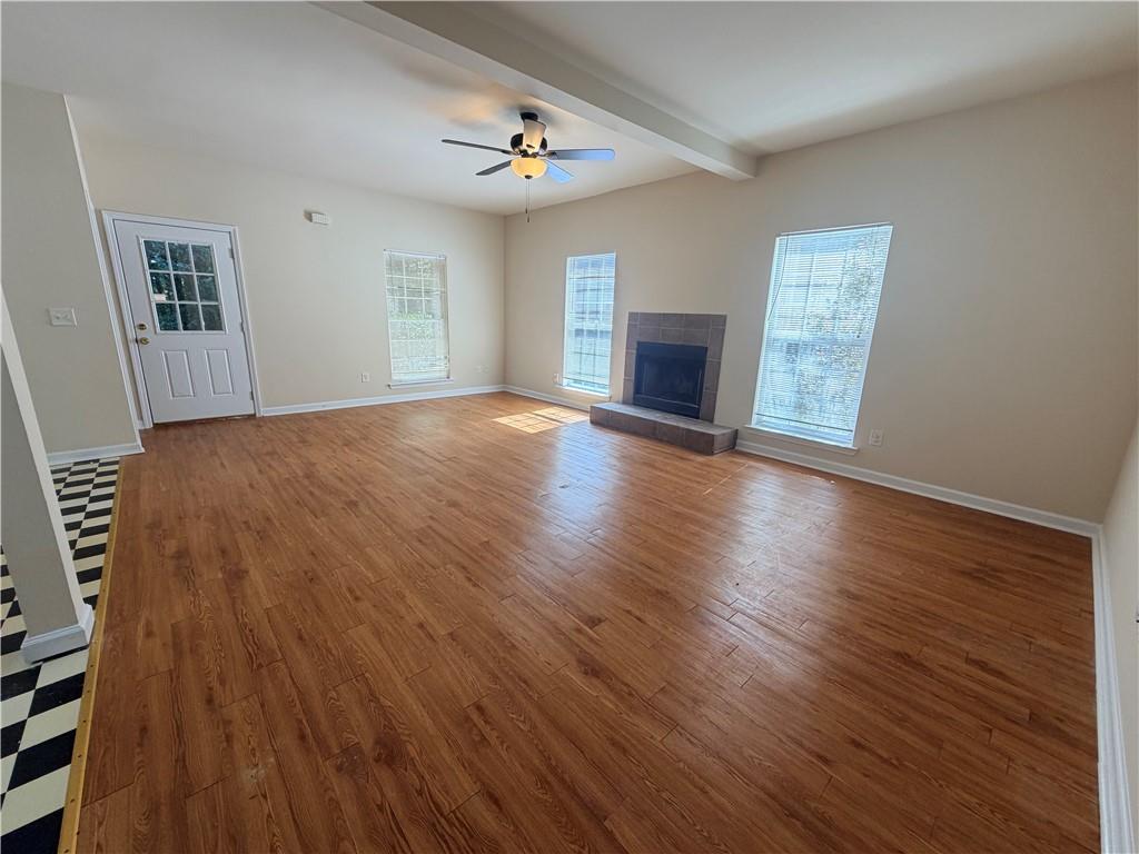 Empty living room with wood floors, a tiled fireplace, and windows.