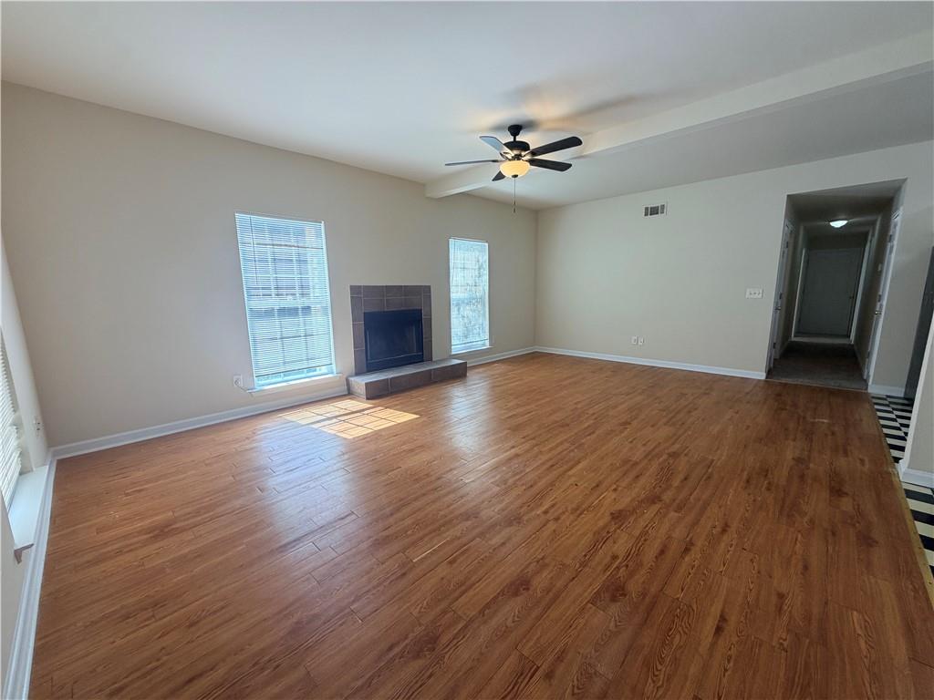 Empty living room with wood floors, a tiled fireplace, two windows, and a ceiling fan.