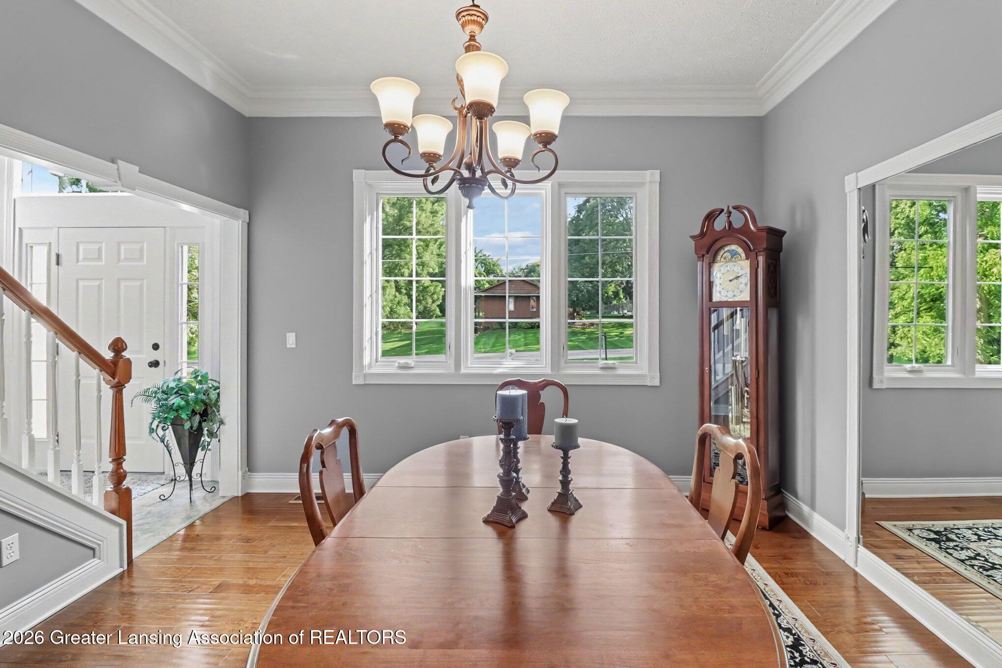 Dining room with a long wooden table, chandelier, and grandfather clock beside windows overlooking a green lawn.