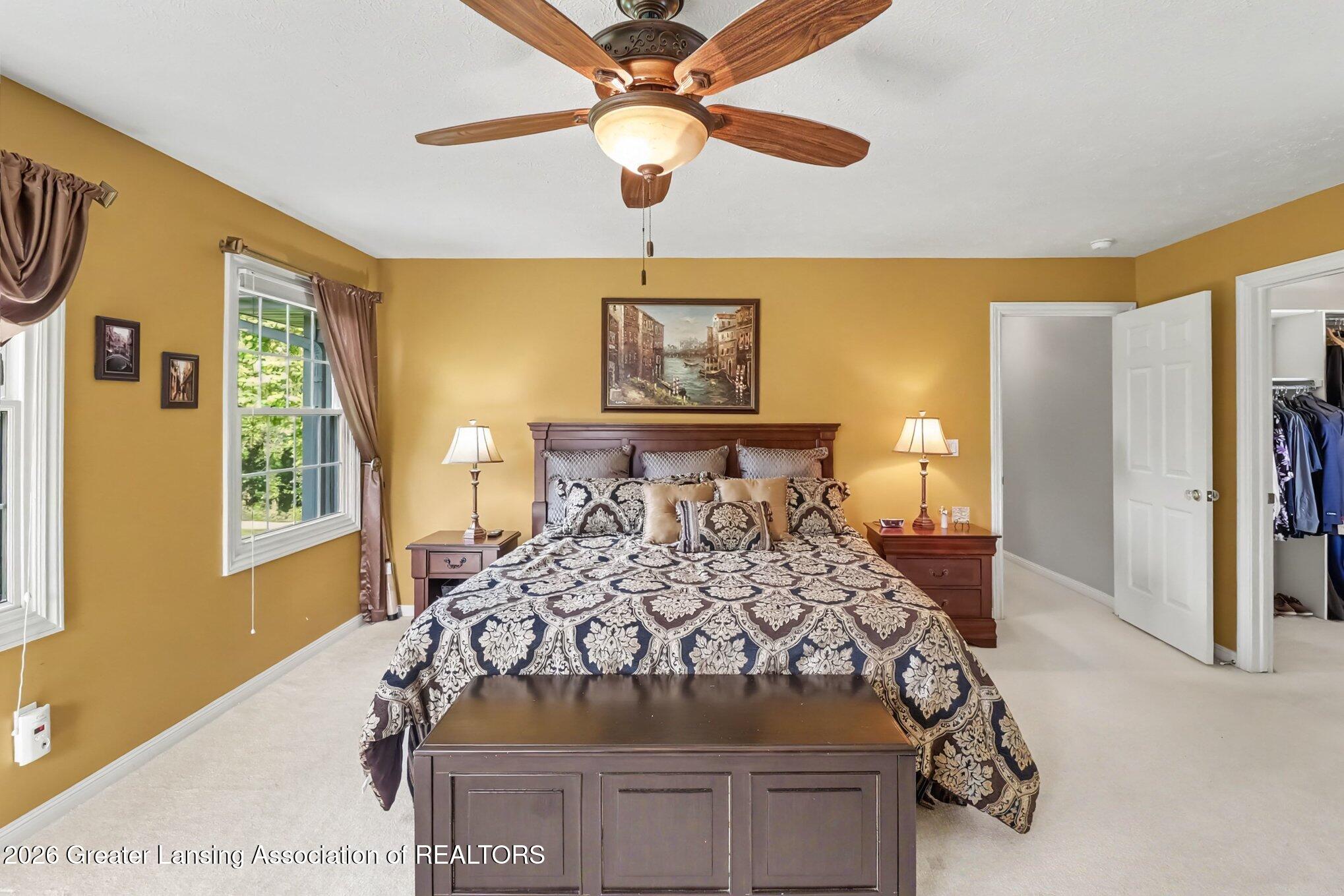 Furnished bedroom with a patterned bed, mustard-yellow walls, and a wooden ceiling fan.