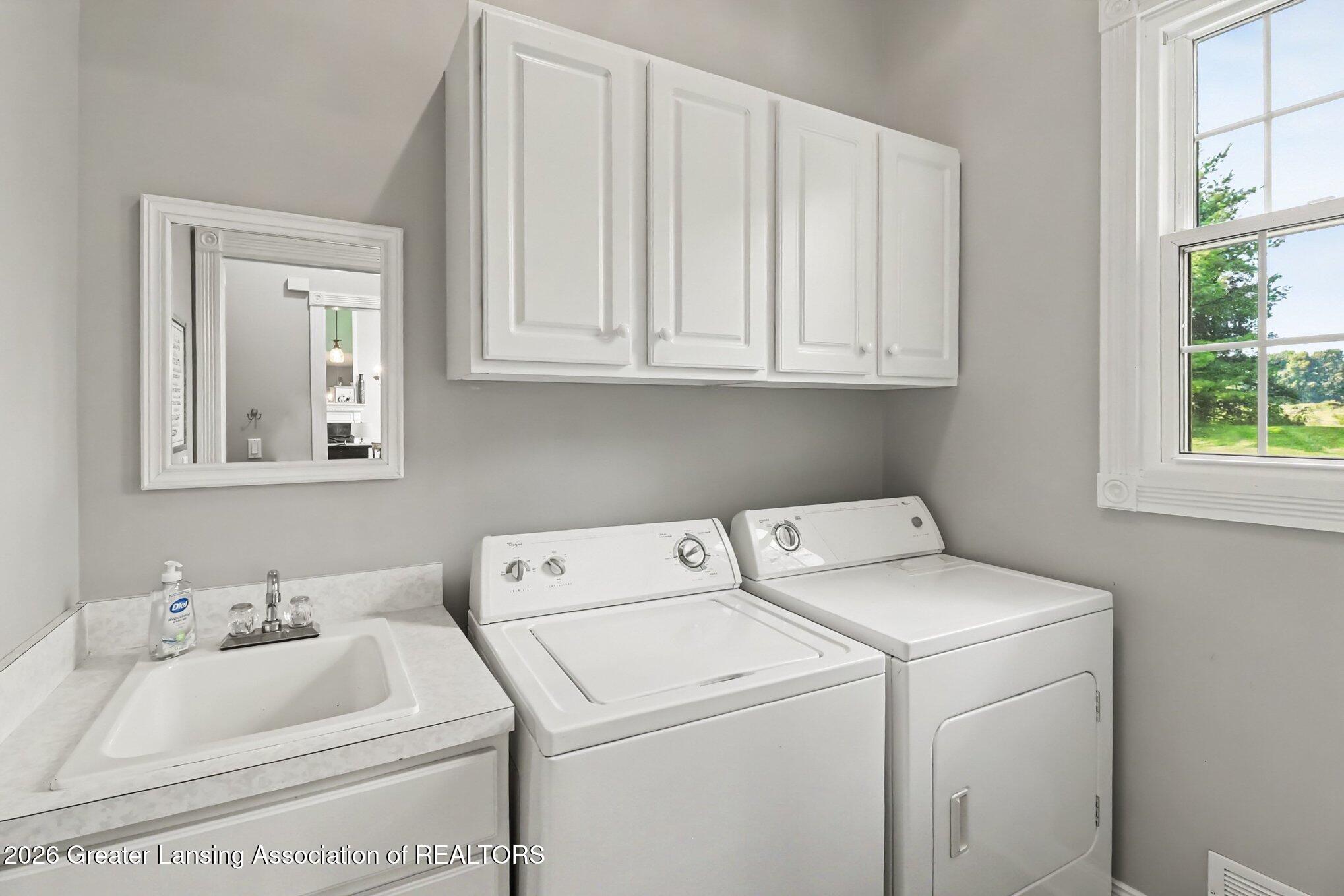 Laundry room with washer, dryer, utility sink, and white cabinets beside a large window.