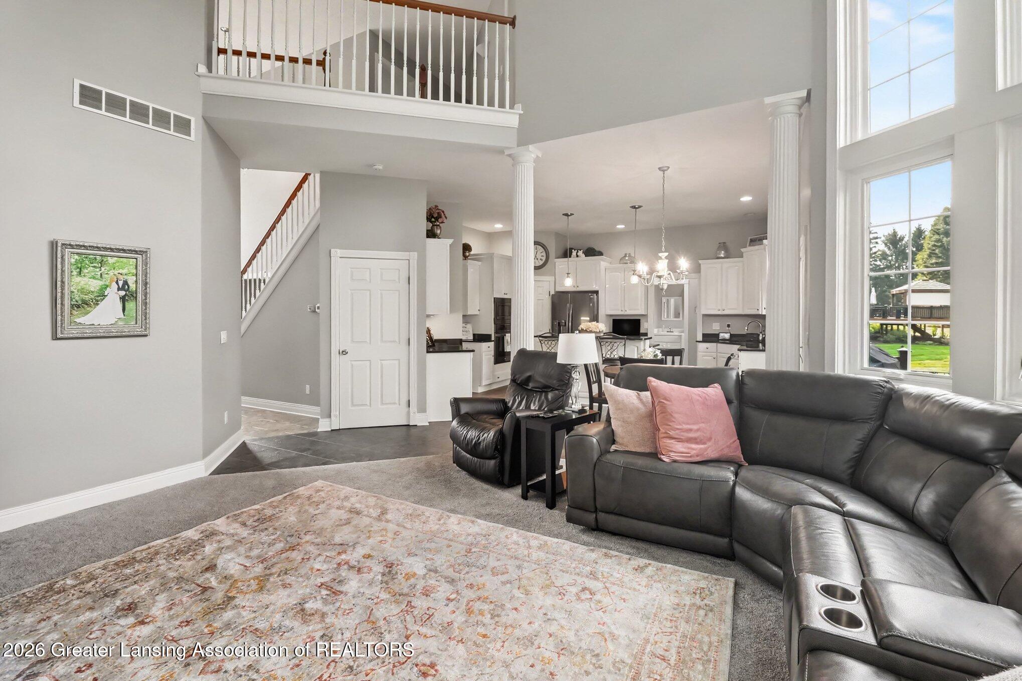 Living room with a dark leather sectional, tall windows, and an open kitchen beneath an upper-level railing.